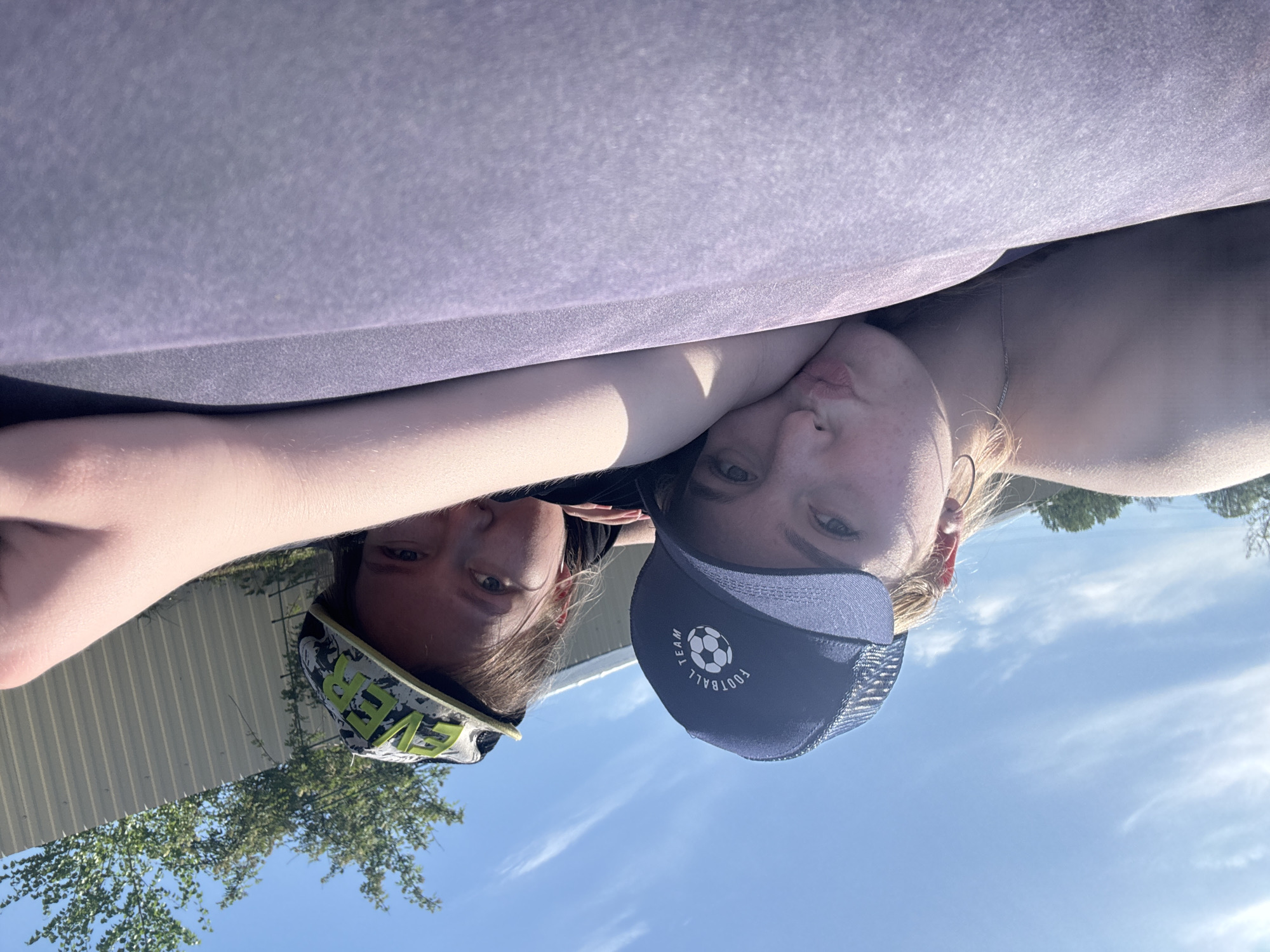 Two individuals, likely young females, are positioned closely together in an outdoor setting under a clear blue sky with scattered cirrus clouds. They appear to be lying down on a surface covered by a purple material. One individual is wearing a dark blue baseball cap with a white soccer ball and the word "FOOTBALL" on it. The other is wearing a baseball cap with the word "EVER" on it in green letters. A metal fence and trees are visible in the background. This is not location identifiable within Zhigalki, Russia.