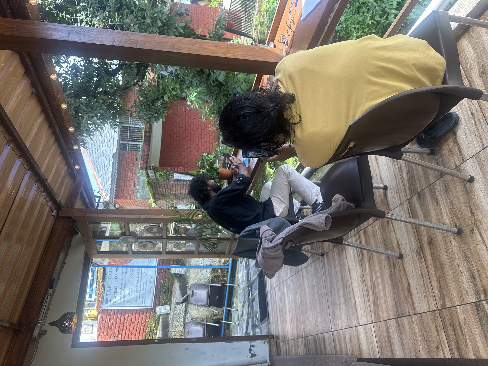 The image depicts an outdoor seating area, possibly a cafe or guesthouse, with a rustic wooden roof and flooring. Two individuals are seated at a wooden counter. The person on the left, a man with curly dark hair, is wearing a black shirt and light-colored pants, and is holding a phone. The person on the right, facing away from the camera, is wearing a yellow t-shirt and is also holding a phone, seemingly taking a picture. In the background, a brick building is visible, with greenery and trees surrounding the area. A sign for "TARU YOGA" is partially visible on the left. The overall impression is a relaxed, outdoor setting, likely in a scenic location. Given the presence of "TARU YOGA", it is plausible this location is associated with yoga or wellness, and the style of the building and surrounding greenery suggests a location in a hilly or mountainous region, consistent with areas like Himachal Pradesh, where places with "TARU YOGA" might be found. However, without more specific visual cues or context, pinpointing an exact location in Haini, India, is not possible solely from the image.