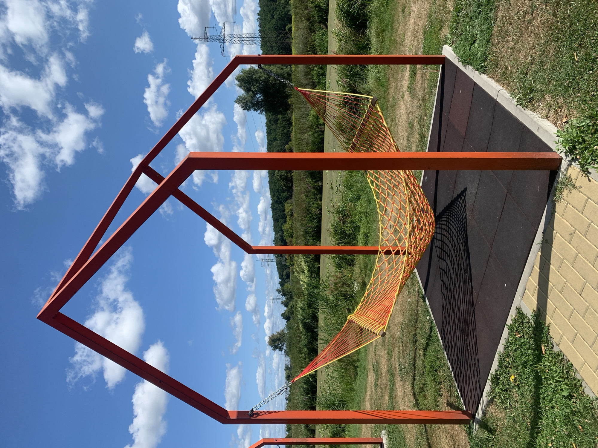 In Irpin, Ukraine, a vibrant orange A-frame structure stands in an outdoor park, supporting a brightly colored orange and yellow net hammock. The hammock is suspended by chains, casting a patterned shadow on the dark rubber matting beneath it. The park setting is characterized by a grassy area with patches of dry grass and some green weeds, bordered by a light yellow brick pathway. In the background, a dense line of trees forms the horizon, with tall electricity pylons visible against a bright blue sky dotted with fluffy white clouds. The scene captures a moment of leisure in a natural, open-air environment.