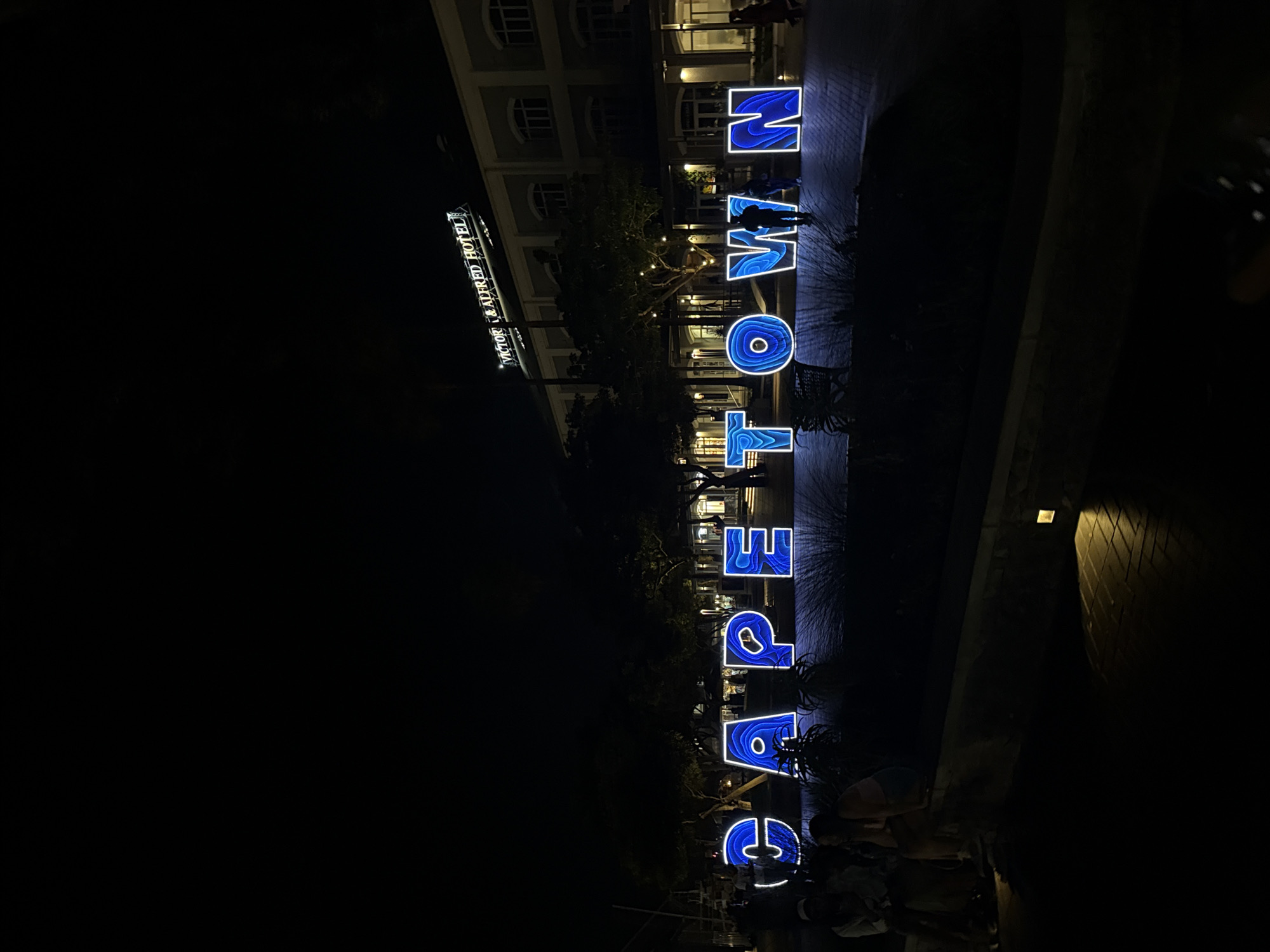 At night, the illuminated vertical sign "CAPE TOWN" is prominently displayed, with each letter rendered in a blue glowing design. The sign stands on a paved area near a building, and people are present near the base of the sign. The "VICTOR & ALFRED HOTEL" sign is also visible in the background. The image captures a public space in Cape Town, South Africa.