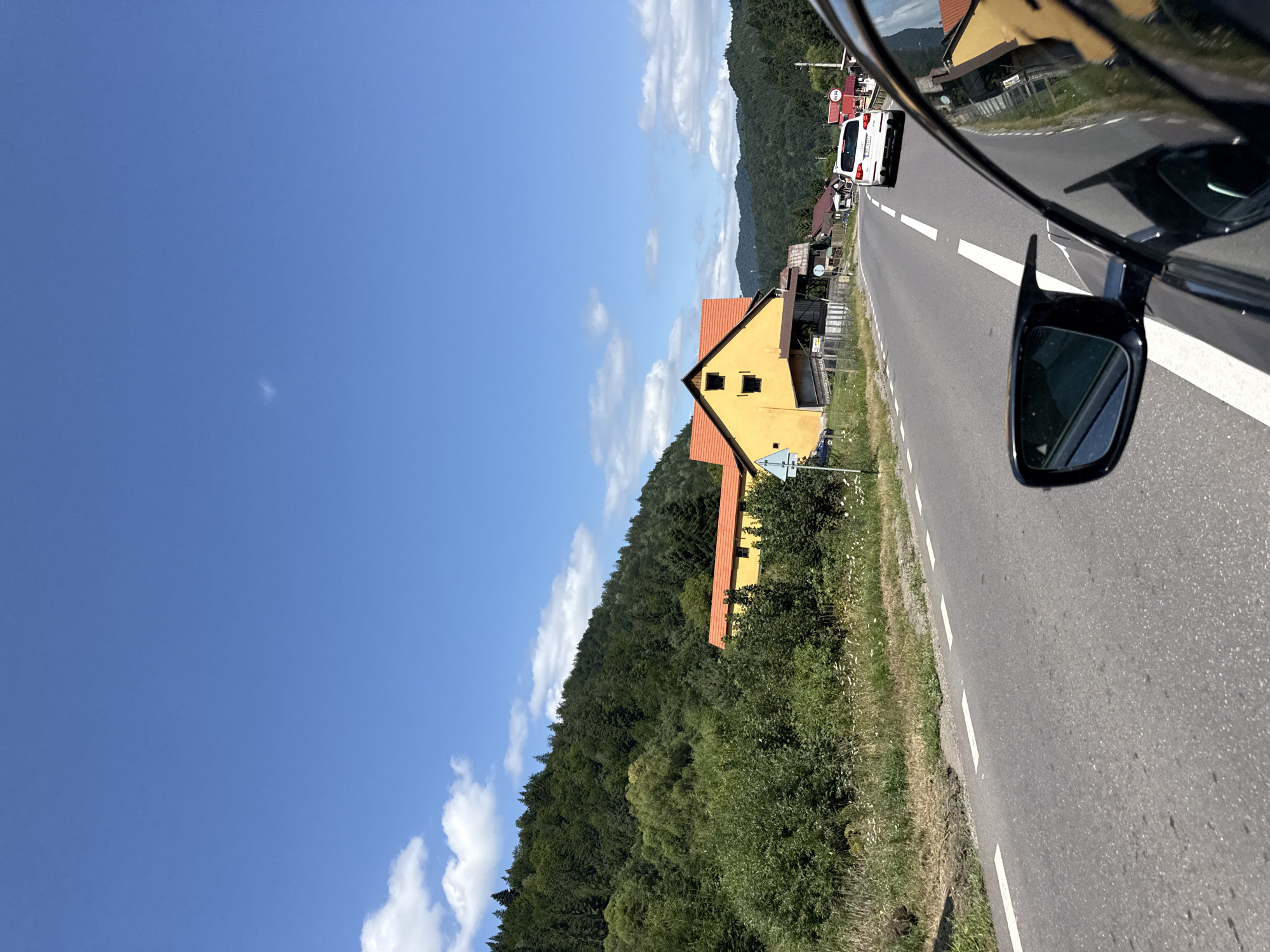 This image captures a view from a car driving along a road in Oituz, Romania. The road is paved with asphalt and has a white dashed line marking the lanes. On the left side of the road, there is a grassy embankment with wildflowers and dense green bushes leading up to a forested hillside. A prominent yellow building with a red-tiled roof is visible on the left, situated on a slight incline. The building has two dark rectangular windows on its upper story and appears to have some sheds or outbuildings attached to it. Further down the road, several other houses with red roofs can be seen, interspersed with trees. A white SUV is driving ahead on the right lane of the road. The sky is a clear blue with a few scattered white clouds. The overall scene depicts a rural roadside environment with residential buildings nestled amidst lush greenery and rolling hills under a bright sky.