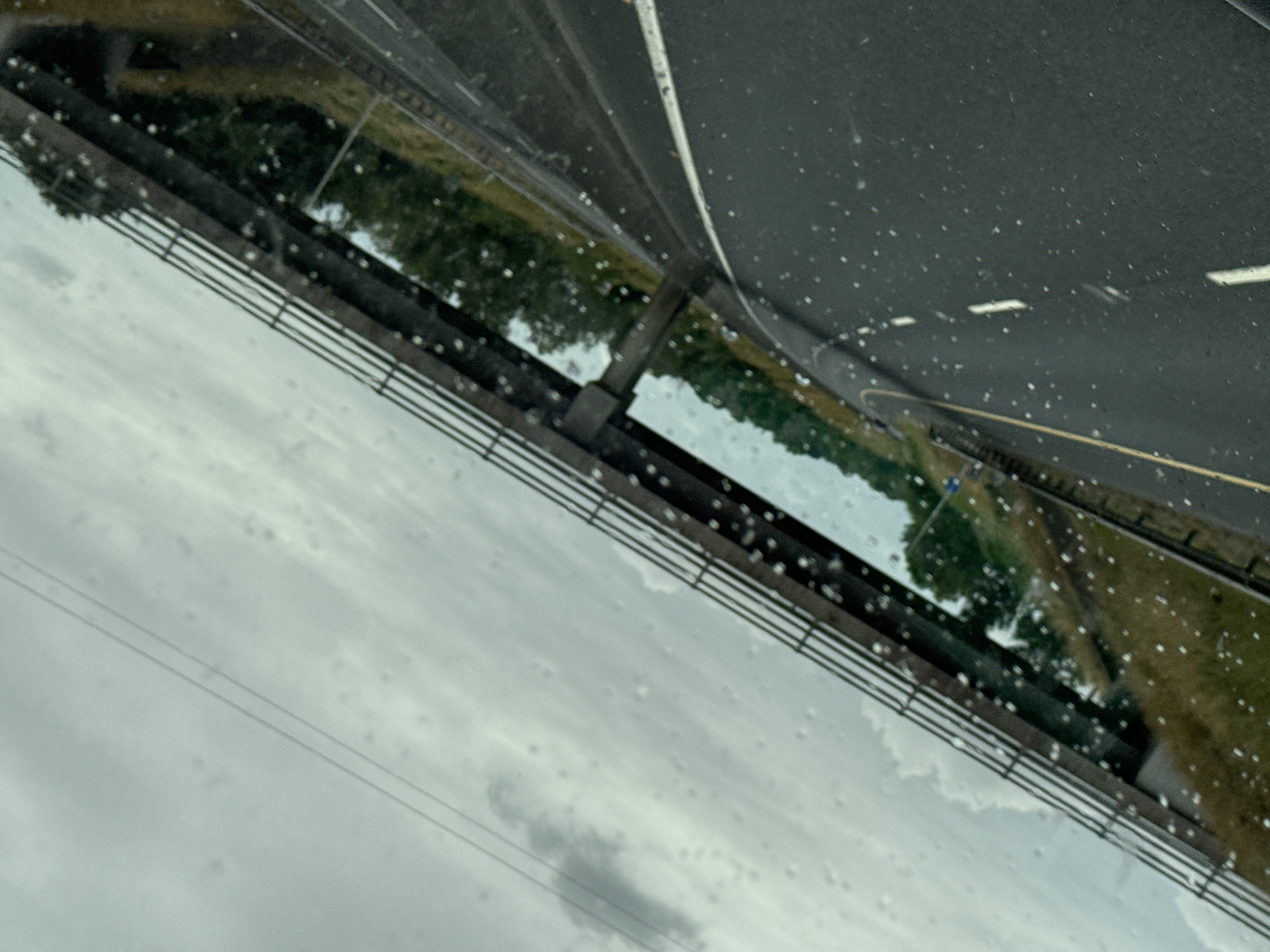 The image, captured from a moving vehicle on a highway, shows a view through a rain-streaked windshield. A concrete overpass, with a metal railing along its edges, spans across the road. The sky is overcast with scattered clouds. The road itself is asphalt, with yellow and white lane markings visible. The surrounding environment consists of green vegetation and trees. The image appears to be taken on the N25 road near Crookstown, County Cork, Ireland.