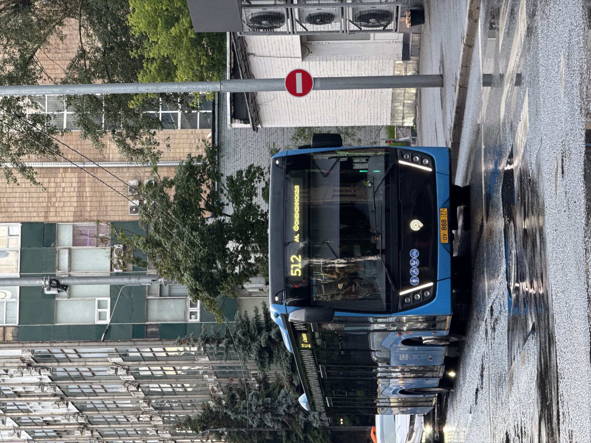 A blue bus with the number 512 and text "м. Фонвизинская" illuminated in yellow on its front is driving on a wet street in Moscow, Russia. The bus has several windows on its side and its headlights are on. A white car is visible behind the bus. The street is wet, indicating it has recently rained, with reflections of the bus and other objects on the wet asphalt. To the right of the bus, there is a white building with air conditioning units installed on its side. A red and white circular "no entry" sign is mounted on a pole next to the building. In the background, there is a multi-story building with numerous windows and a brick-clad building. Trees are present on the left side of the street and between the buildings.