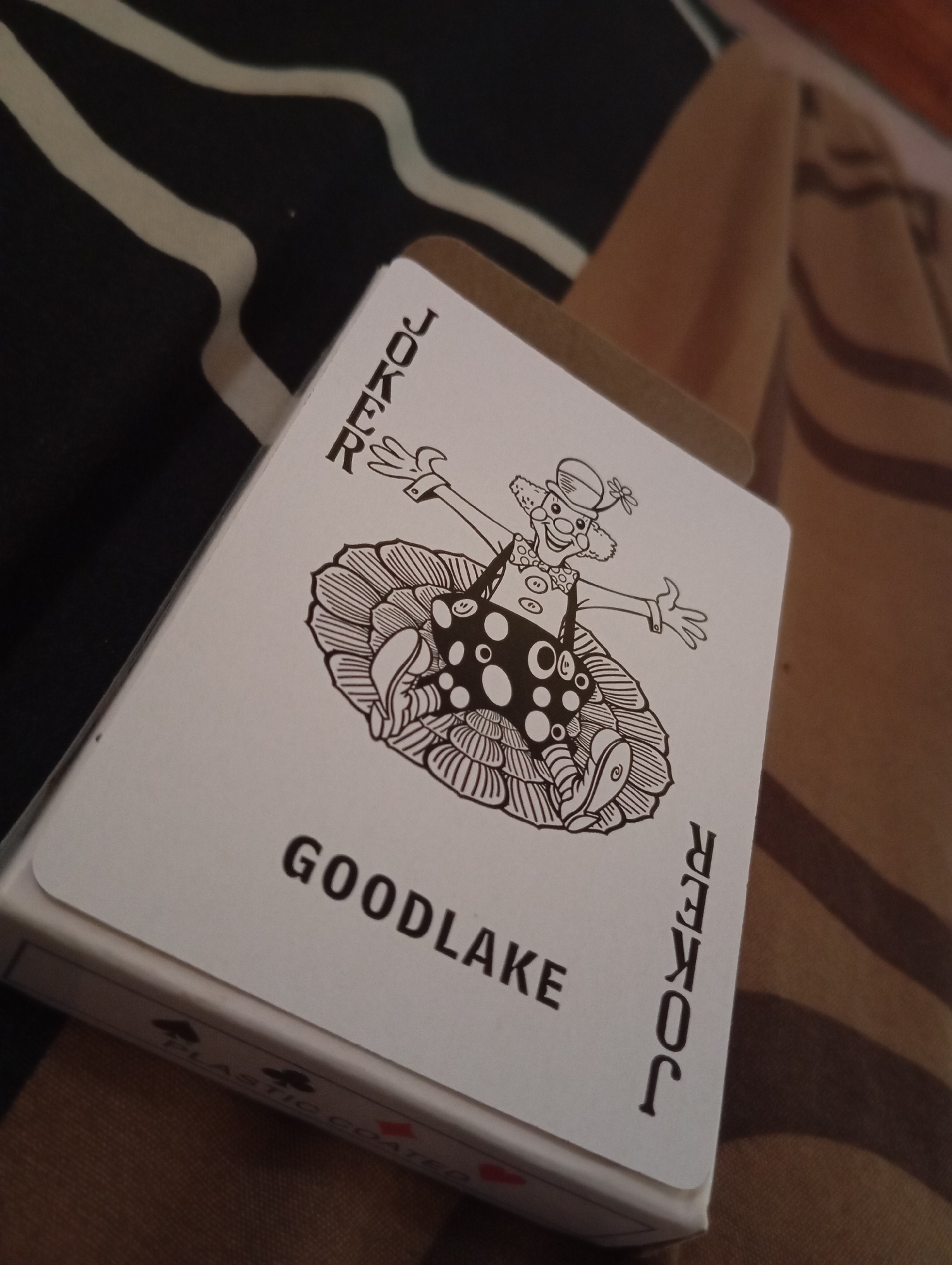 A close-up, low-angle shot shows a white-faced joker playing card from a Goodlake deck. The card is angled diagonally from the bottom left to the top right, positioned on a surface with a striped pattern of black and off-white, and partially on a brown textured fabric. The joker illustration depicts a clown with a smiling face, wearing a hat with a flower, and a suit with polka dots. The word "JOKER" is printed vertically along the left and right edges of the card. The bottom edge of the card box is visible, showing the words "PLASTIC COATED" and small suit symbols. The background consists of folded brown fabric, likely bedding or clothing, which is out of focus. The lighting is dim and directional, casting shadows and highlighting the texture of the card and fabric. The scene appears to be indoors.