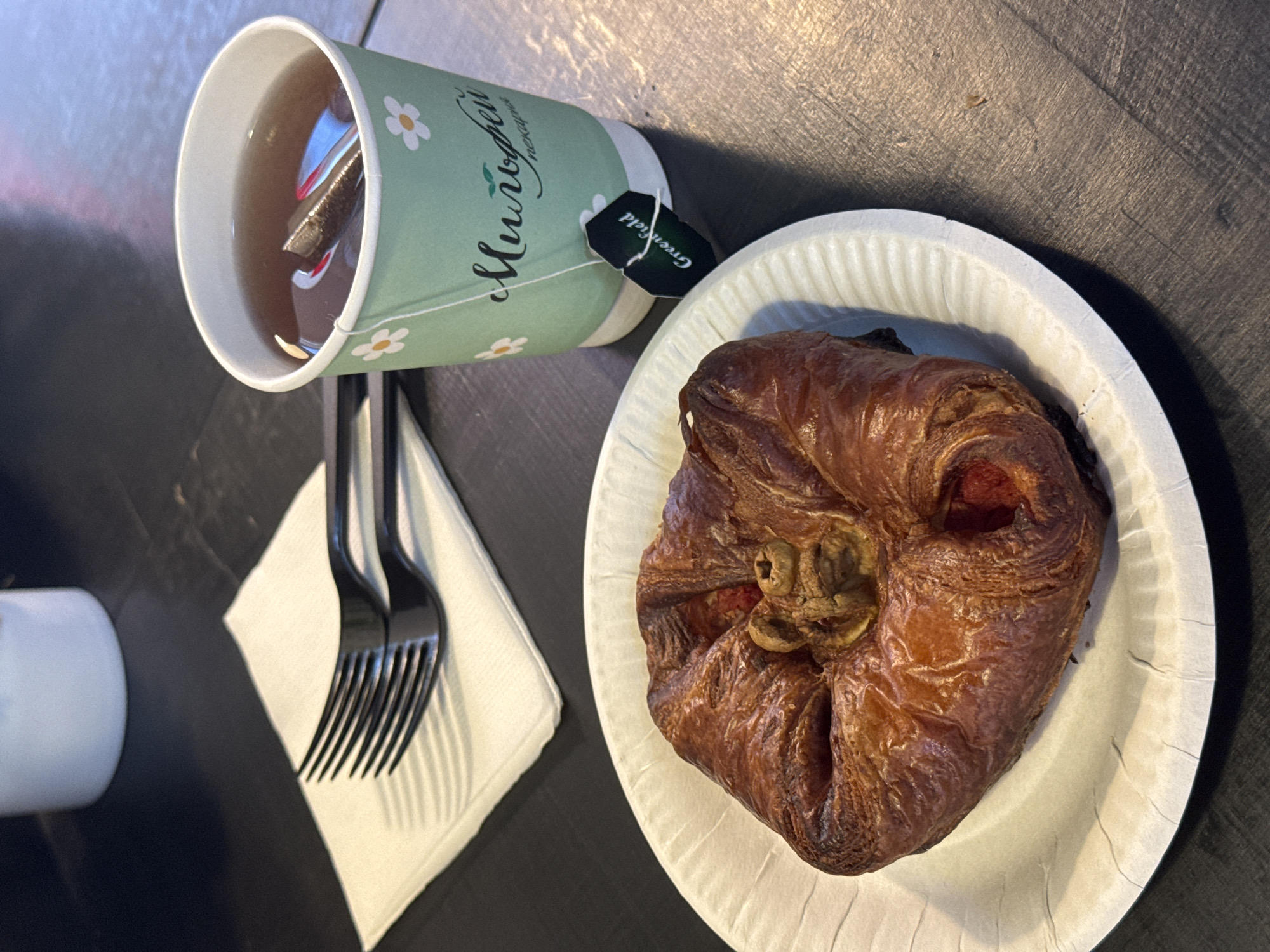 A Danish pastry sits on a white paper plate, placed on a dark wooden table. Next to the plate, two black plastic forks are stacked on a white napkin. To the right of the napkin, a light green paper cup filled with tea is visible. A tea bag tag, labeled "Greenfield," hangs over the rim of the cup. The cup has a daisy pattern and Russian text that translates to "Milyufey Bakery." The background shows a blurry white cup and a dark surface.