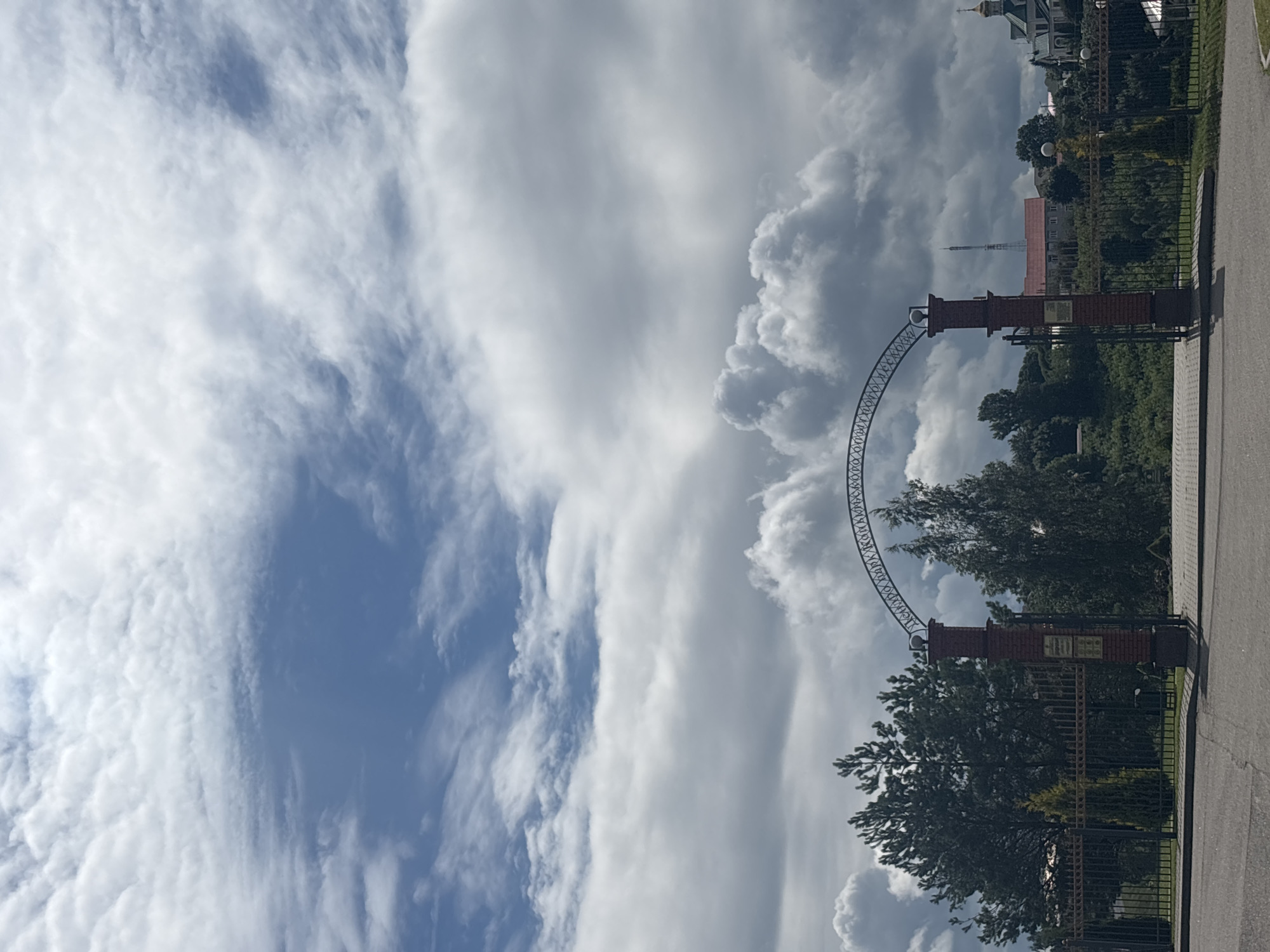 The image shows an outdoor scene in Kapyĺ, Belarus, on a partly cloudy day. The camera is oriented vertically. In the foreground, there is a paved pathway and a black metal fence. Behind the fence, there are trees and a brick archway structure. The archway has an ornate metal frame connecting two brick pillars. A sign is attached to one of the pillars. In the background, more trees and buildings are visible, including a church with a golden dome and a tall, slender tower. The sky is filled with a mix of white and gray clouds against a blue sky, suggesting it is daytime. The overall impression is that of an entrance to a park or a historical site.