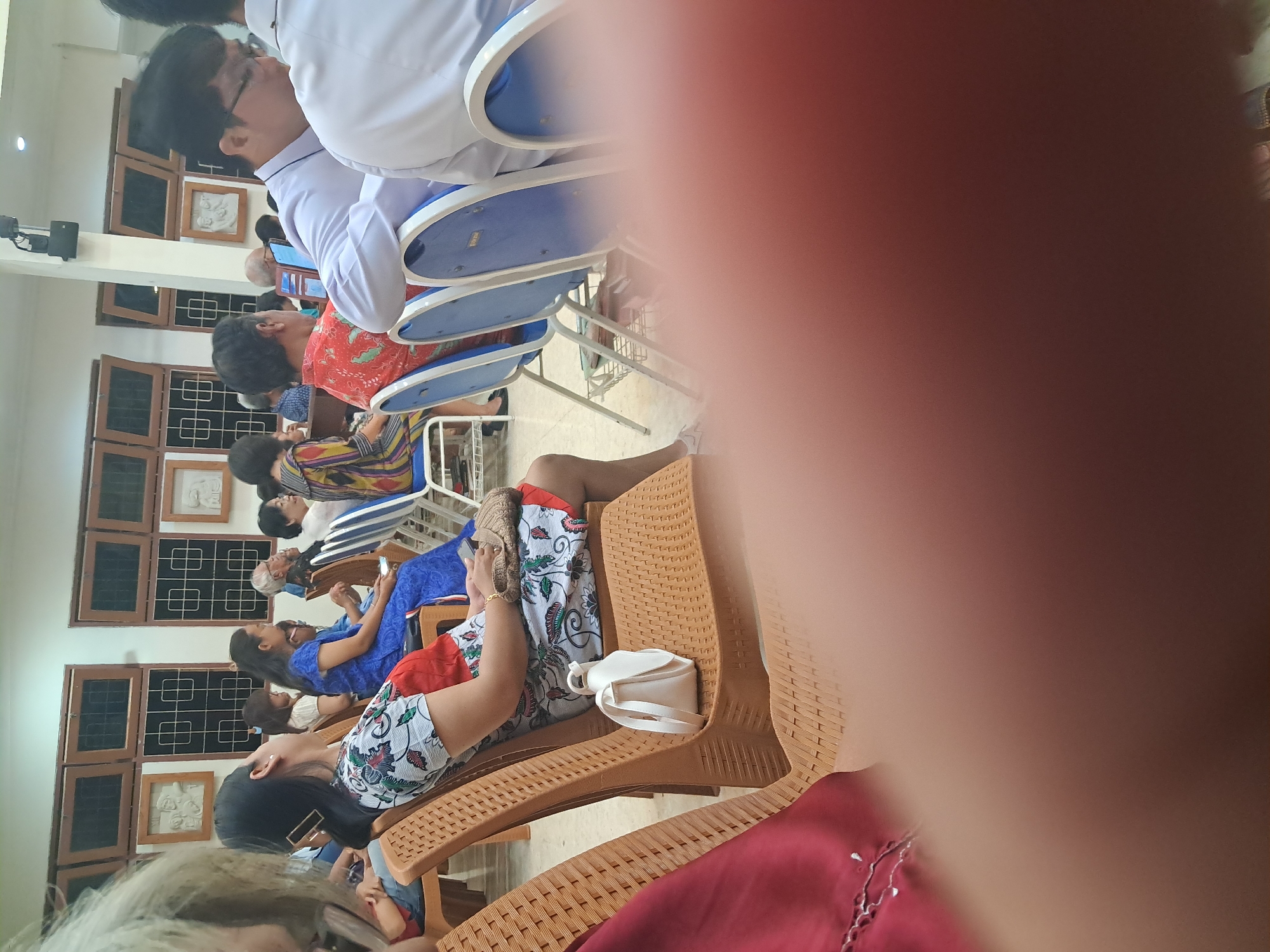 The image shows an indoor gathering of people, likely at a religious or community event, in what appears to be a hall in Ngaglik, Indonesia. In the foreground, out of focus, a person's hand and chest are visible, draped in red fabric. Seated in rows of chairs are several individuals, mostly facing forward. One woman in a patterned dress is seated in the center, looking upwards with her eyes closed. Next to her, a woman in a blue dress is seated. Further back, individuals are visible in various attire, including a man in a white shirt and glasses, and others wearing colorful clothing. The hall features wooden-framed windows with dark panes and decorative wall art. The chairs are made of brown plastic with woven-like textures, and some have blue seats. The lighting suggests an indoor setting with artificial illumination.