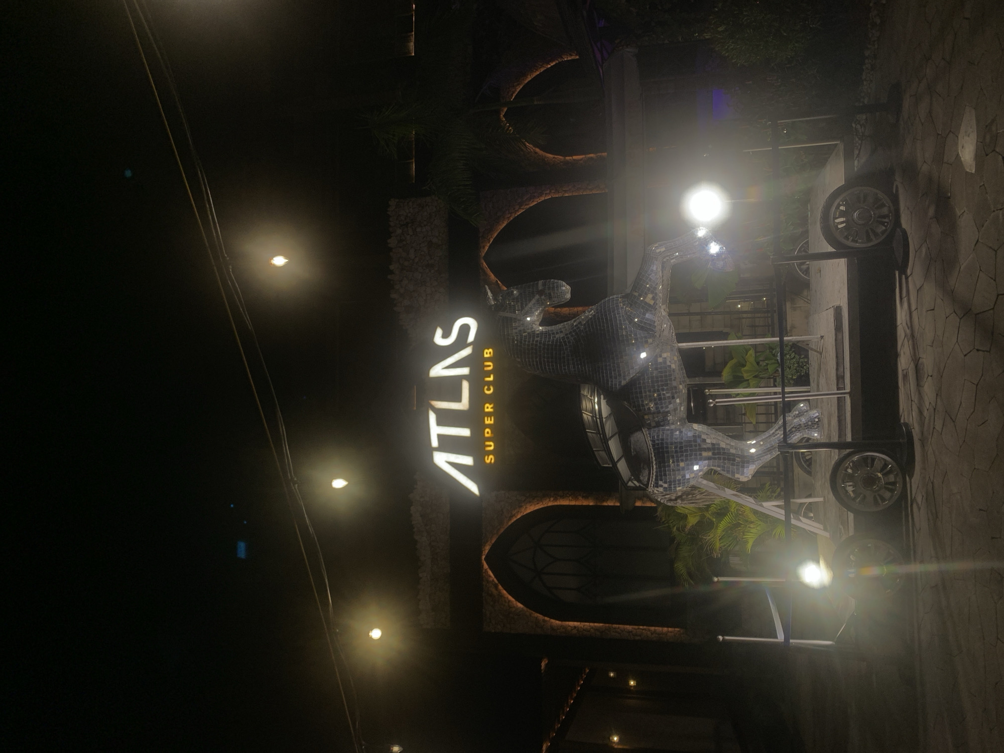 The image depicts the illuminated entrance area of "ATLAS SUPER CLUB" at night, located in North Kuta, Indonesia. The dominant feature is a large, brightly lit sign displaying "ATLAS" in prominent white letters, with "SUPER CLUB" written in smaller text below.
In front of the sign, to its right, stands a striking, life-sized sculpture of an animal, possibly a deer or stag, entirely covered in reflective mirror tiles. This sculpture gleams under the artificial light, creating numerous sparkling reflections. The club's facade features a textured stone-like finish with distinct arched architectural elements, including dark, ornate windows or doorways.
Various palm trees and other foliage are visible behind and around the main structure, suggesting a tropical environment. The ground is paved with irregular stone slabs. Several bright artificial lights illuminate the scene, some creating noticeable lens flares. On the far right, part of a wheeled structure, possibly a mobile cart or stand, is visible with a prominent wheel. Overhead, utility lines or cables crisscross against the dark night sky. No people are visible, but the bright lighting and the nature of the establishment suggest an active or soon-to-be active nightlife venue. The atmosphere conveyed is one of vibrant entertainment and readiness for guests.