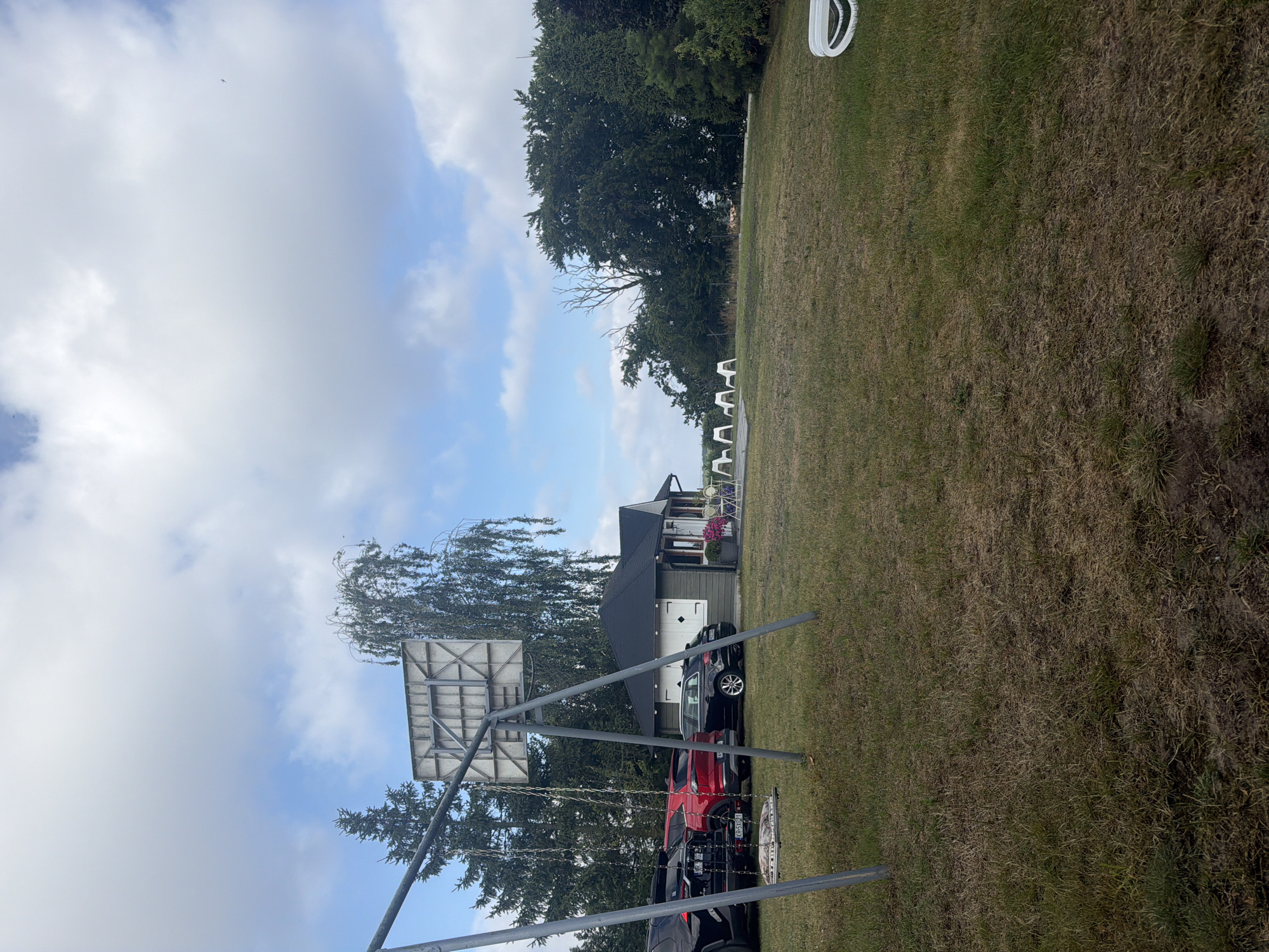 The image depicts an outdoor scene in Międzyzdroje, Poland. In the foreground, a metal swing set with a single swing is visible on a grassy area. Behind the swing set, several cars are parked, including a black SUV, a red SUV, and a black sedan. A small, dark-colored building with a peaked roof and white trim is situated behind the cars. In front of the building, there are several white chairs arranged around a table. Further in the background, a dense line of trees forms a backdrop under a cloudy sky. The overall impression is that of a residential or recreational area.