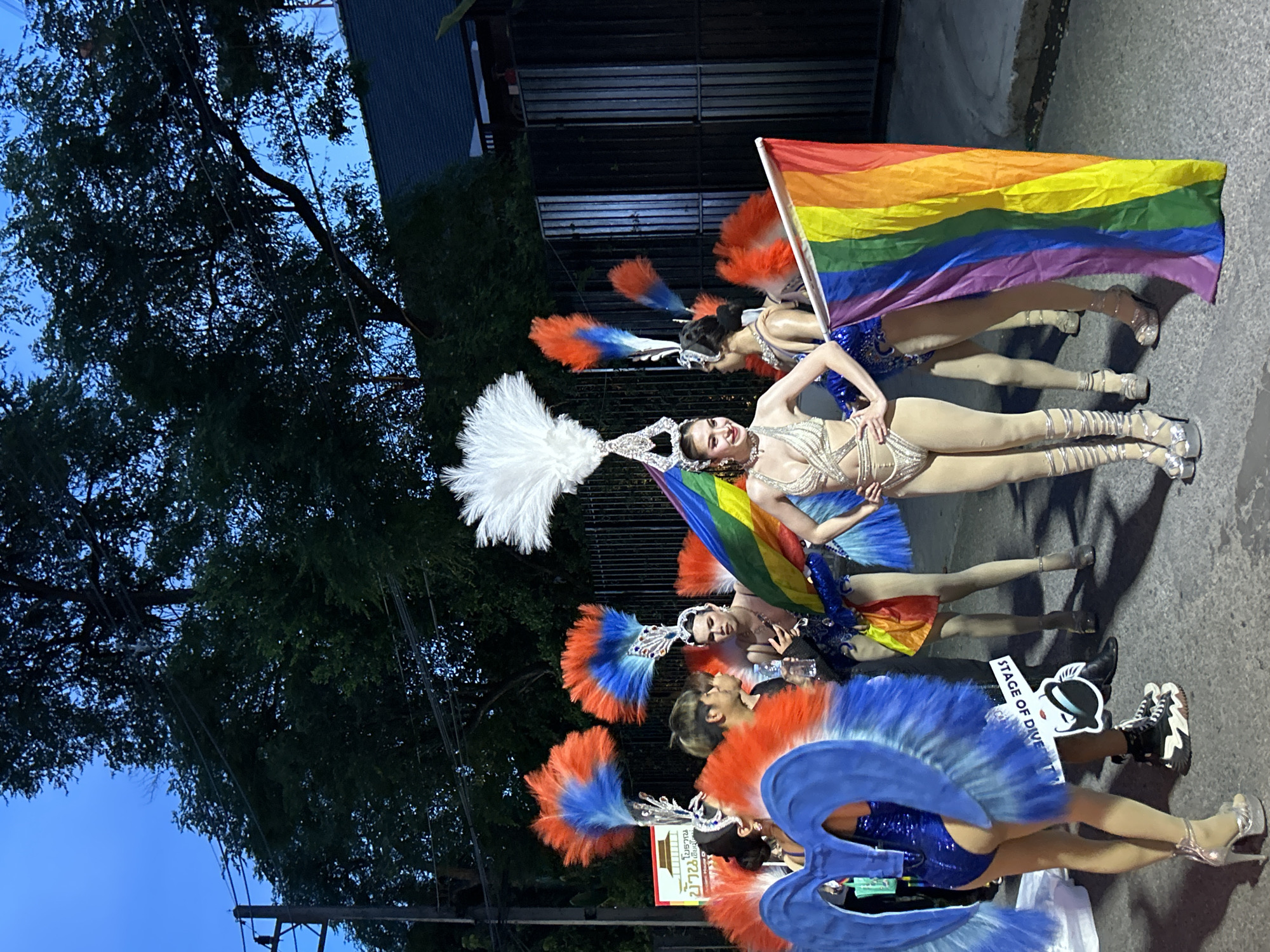 A group of people in elaborate costumes are posing in front of a building. The costumes are colorful, with feathers and sequins. A rainbow flag is also visible. The sign reads "Stage of Diversity". The location is likely Chiang Mai City Municipality, Thailand.
