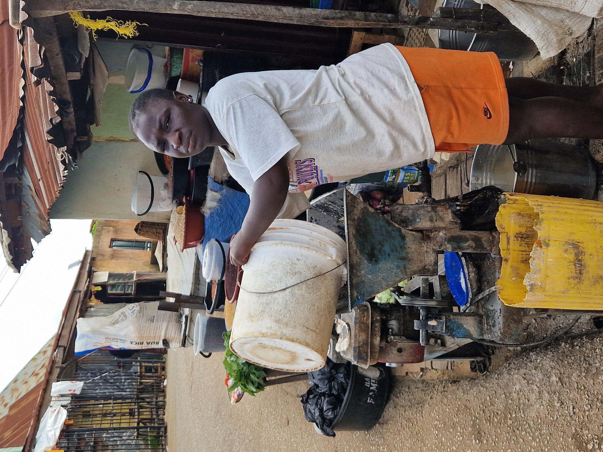 A Black woman, identified as an adult, stands outdoors in what appears to be a market setting. She is wearing a light-colored t-shirt with colorful lettering and orange shorts. The woman is in the process of pouring liquid from a large, white bucket into a blue, funnel-like apparatus that sits atop a rusty machine. Bags of black material are visible in a black container to her left, and a yellow plastic container is in the foreground to her right. Several buckets of varying sizes and materials, some filled with what appear to be grains or dry goods, are also present around her. In the background, a building with a corrugated metal roof and walls made of varied materials is visible, along with sacks of rice and other market stalls. The ground is a mixture of dirt and gravel. The overall environment suggests an informal commercial area.