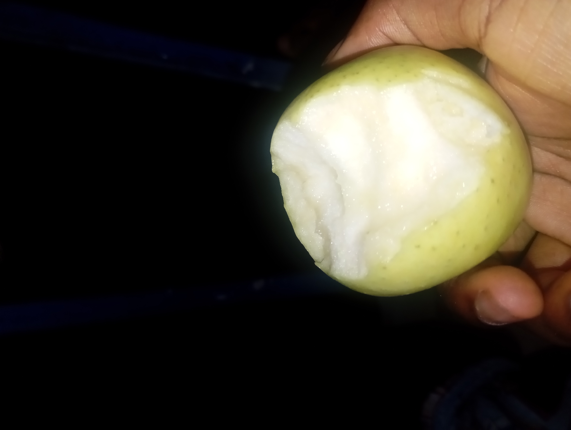 A close-up, slightly blurry image shows a person's hand holding a partially eaten light green apple against a dark, out-of-focus background. The apple is roughly spherical and has a noticeable bite taken out of its side, revealing the white, fibrous flesh. The skin of the apple is smooth with small, dark speckles. The hand holding the apple appears to be an adult's, with dark skin. The background consists of deep shadows and indistinct shapes, suggesting an indoor or dimly lit environment.