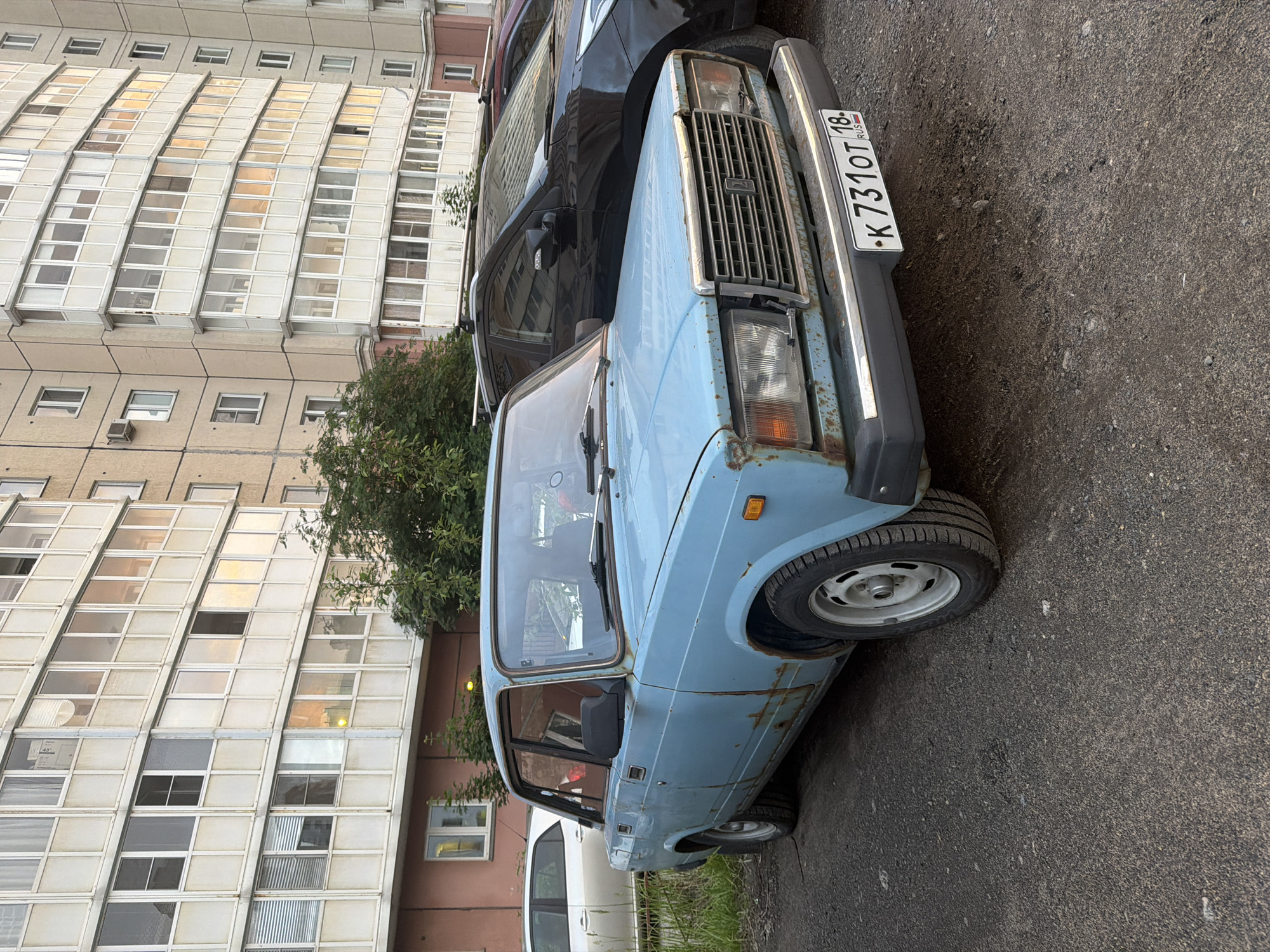 In the city of Unknown, Unknown, a light blue, heavily rusted sedan is parked on a gravel surface in front of a modern apartment building. The car, a Lada, is weathered and shows significant signs of decay, particularly around the wheel wells and door panels. To the right of the Lada, a dark-colored SUV is partially visible. The apartment building behind the vehicles features a distinctive design with numerous balconies and windows, reflecting the late afternoon or early evening light. Green foliage, including trees and grass, surrounds the parking area. The license plate of the Lada is visible and reads "K 731 OT 18 RUS". There are no people in the frame.