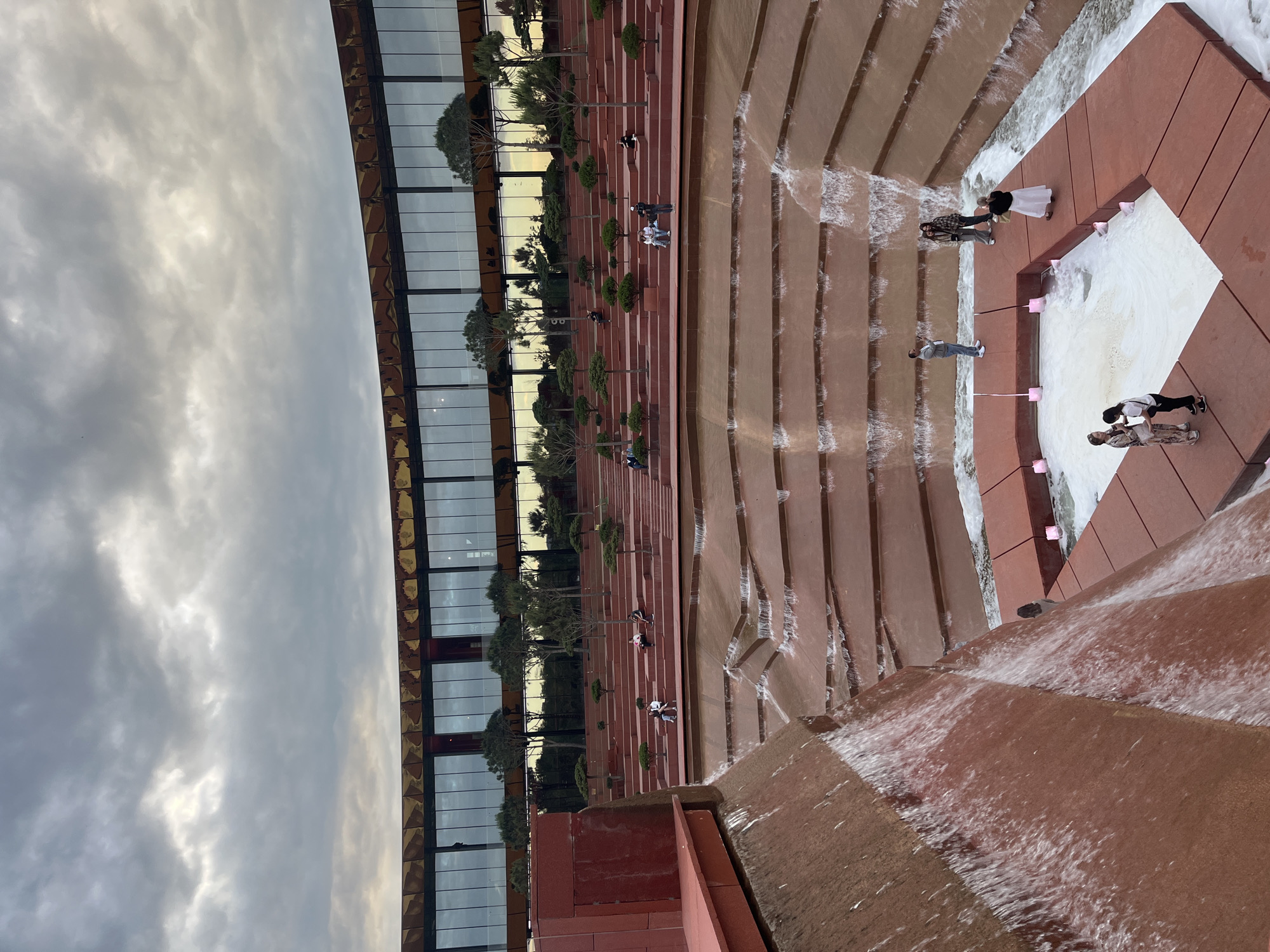 The image depicts a tiered waterfall structure at the Park Krasnodar, Russia. The structure is constructed of red brick. People are dispersed on the stairs of the structure. A modern building with glass windows is behind the waterfall. The sky is cloudy.