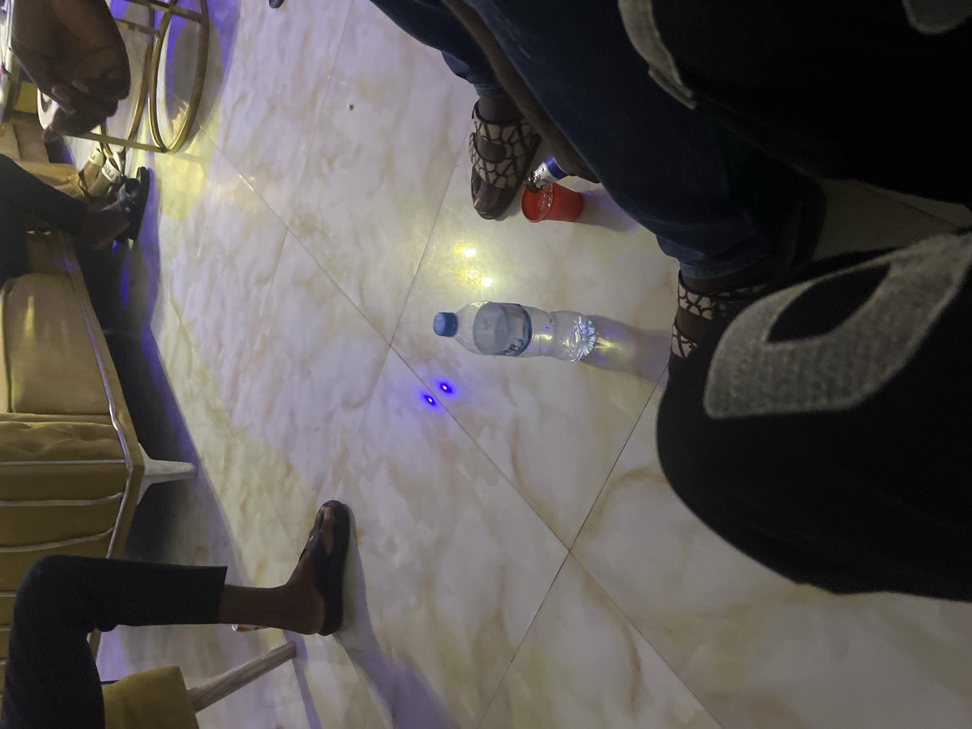 In Jauro Yinu, Nigeria, a group of individuals are gathered around a table, with a focus on their lower extremities and the floor. The floor is tiled with a beige and white marble pattern. In the foreground, a person's legs are visible, clad in dark clothing and patterned sandals. To the right, another person is wearing blue jeans and sandals. A partially visible plastic cup with liquid and a bottle of water are placed on the floor between them. Further back, another individual's legs are seen wearing black pants and black sandals. The scene appears to be indoors, likely in a social setting, with furniture that includes a mustard-yellow couch and a metallic gold-rimmed chair visible in the background. Ambient lighting, including blue and yellow hues, is present.