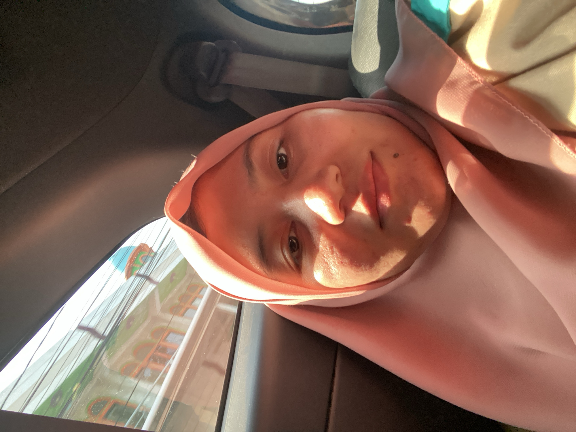 A medium shot selfie shows a young woman wearing a pink hijab, sitting in the back seat of a car. She is looking directly at the camera, with her face partially illuminated by sunlight. In the background, visible through the car window, is a street scene in Antasan Senor Ilir, Indonesia. The scene depicts a building with green and orange accents, possibly a mosque, with a blue dome. Power lines are also visible crisscrossing the sky. The interior of the car shows a dark gray headliner and a gray seatbelt. The woman's clothing appears to be a beige and teal garment under the hijab.