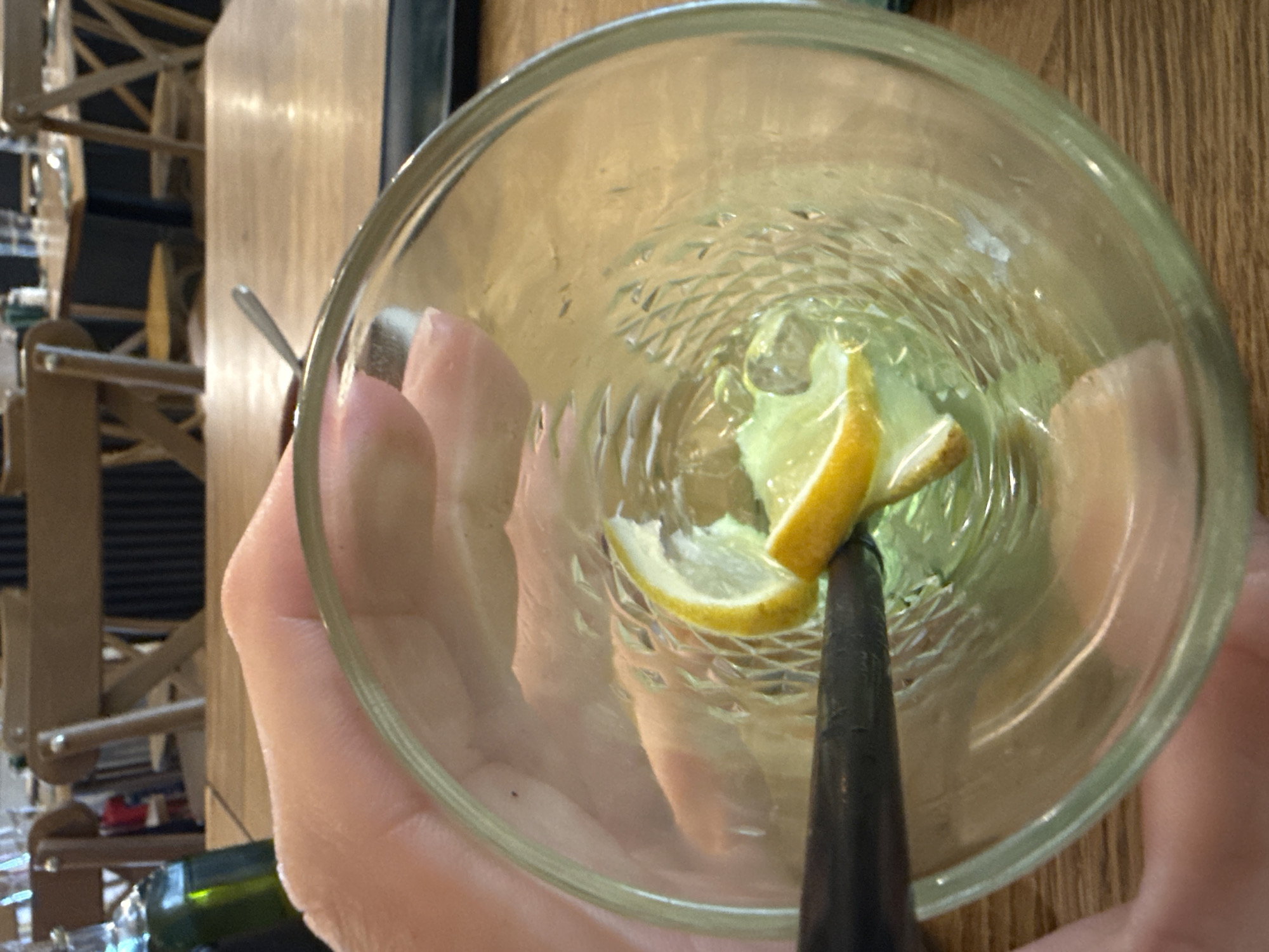 A top-down view shows a clear, patterned glass holding remnants of a drink, including lemon slices, possibly on ice, with a black straw. The glass is being held by a person's hand. The background includes a wooden table surface, a metal spoon, chairs, and a bottle. The location is likely Sofia, Bulgaria, based on context clues not visible in the image.