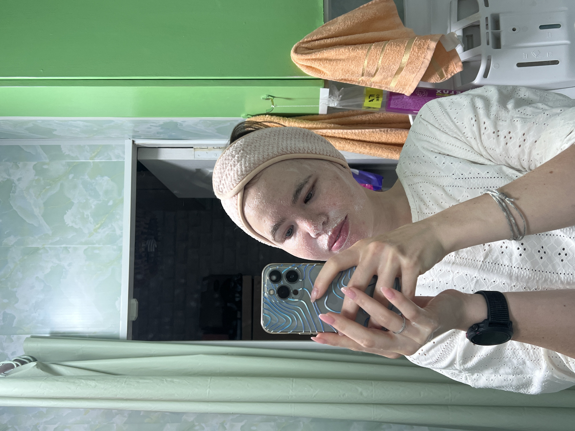 A young woman is captured in a mirror selfie, showing her reflection in what appears to be a bathroom or utility room. The image is oriented vertically, but the woman holds her phone horizontally, making the scene appear rotated.
The woman has light skin, dark hair pulled back into a bun, and wears a cream-colored, textured headband. Her face is covered with a white or foamy face mask, indicating a skincare routine. She is wearing a light-colored, possibly off-white, top with an eyelet lace pattern. On her left wrist, she wears a black digital watch, and on her right, a silver bracelet. A delicate ring is visible on her left ring finger. She holds a silver smartphone, likely an iPhone, with a distinctive blue and white wavy patterned case, using her left hand to frame the selfie.
The background shows various elements of the room. A vibrant green painted surface, possibly cabinetry or a shelf, dominates the upper part of the background. Below this, there's a light-colored, possibly marbled, wall or paneling. An orange or peach-colored towel is draped or hung in the middle-right, behind which a white appliance with grilles and compartments is visible, possibly a washing machine or dryer. Further to the left, behind the towel, a purple container with mirrored text "XUЯOX" and a yellow label with mirrored text "MBO" are partially visible. A light green, possibly fabric or plastic, barrier or curtain is visible in the lower part of the frame.
The woman's expression is neutral and focused on taking the photo, suggesting a routine self-care moment. The setting is indoors, typical of a home environment in Chaikovsky, Russia.