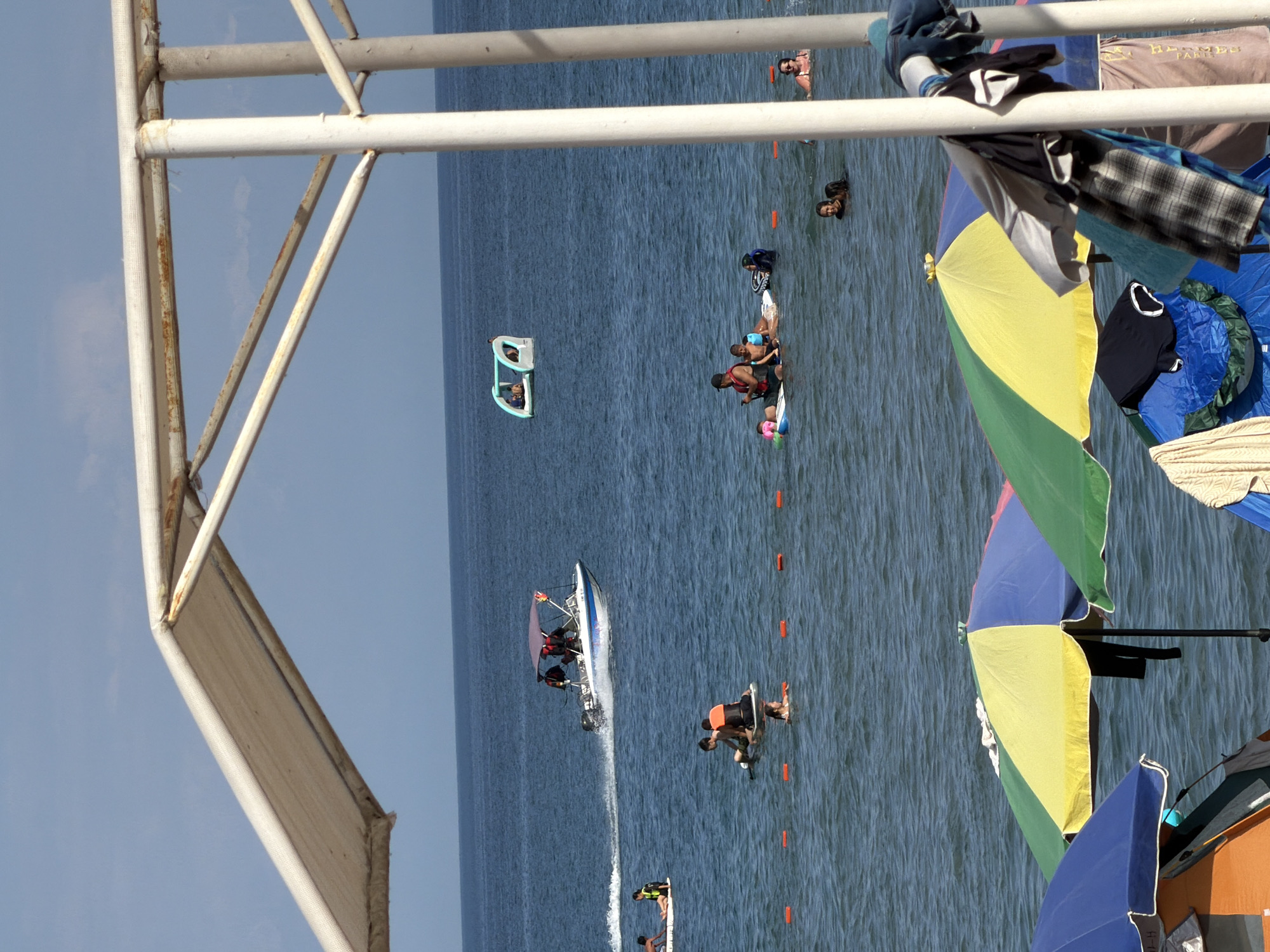 An outdoor daytime scene at a beach shows several people in the water and on the shore. In the foreground, a white canopy structure with weathered metal supports is visible. Colorful beach umbrellas, including a yellow, green, and blue one, are set up on the shore. A boat speeds across the water in the mid-ground, leaving a white wake. Several individuals are seen on inflatable rafts and paddling boards. Another group is swimming in the water. In the background, the clear blue ocean meets a pale blue sky. The overall impression is a lively beach day with various recreational activities.