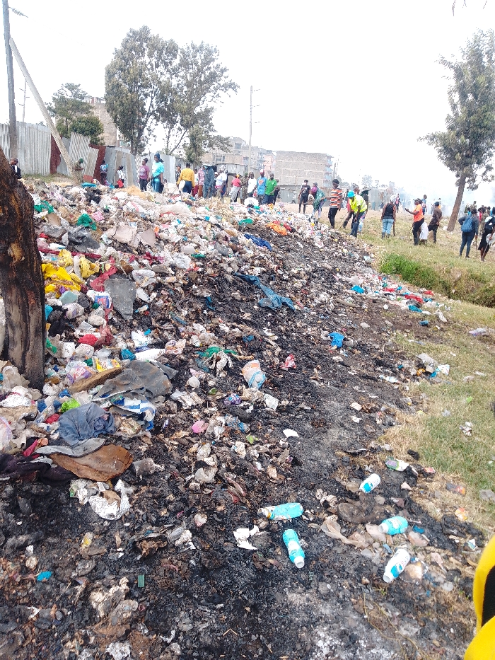 A large pile of garbage dominates the scene, featuring plastic bags, bottles, and other debris. Some parts appear to be burned, with black, charred areas visible. A group of people is visible in the background, standing near the edge of the dump. The setting appears to be an urban or suburban area with buildings and trees in the distance. The overall scene depicts environmental pollution and a potential health hazard.