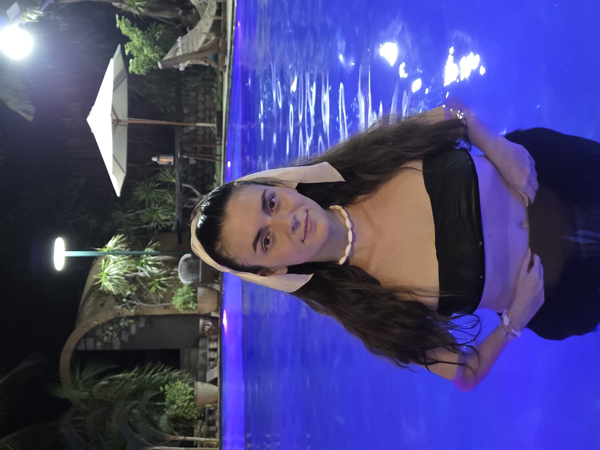 A woman is partially submerged in a brightly illuminated pool, presumably at night. She has long brown hair, a white headband, and is wearing a black top and a seashell necklace. A white bracelet is visible on her right wrist. The pool water is a vivid blue, reflecting the light. In the background, there's a building with plants and a street lamp, along with a white umbrella. The scene appears to be at a resort in Mũi Né, Vietnam, given the tropical vegetation and architecture.