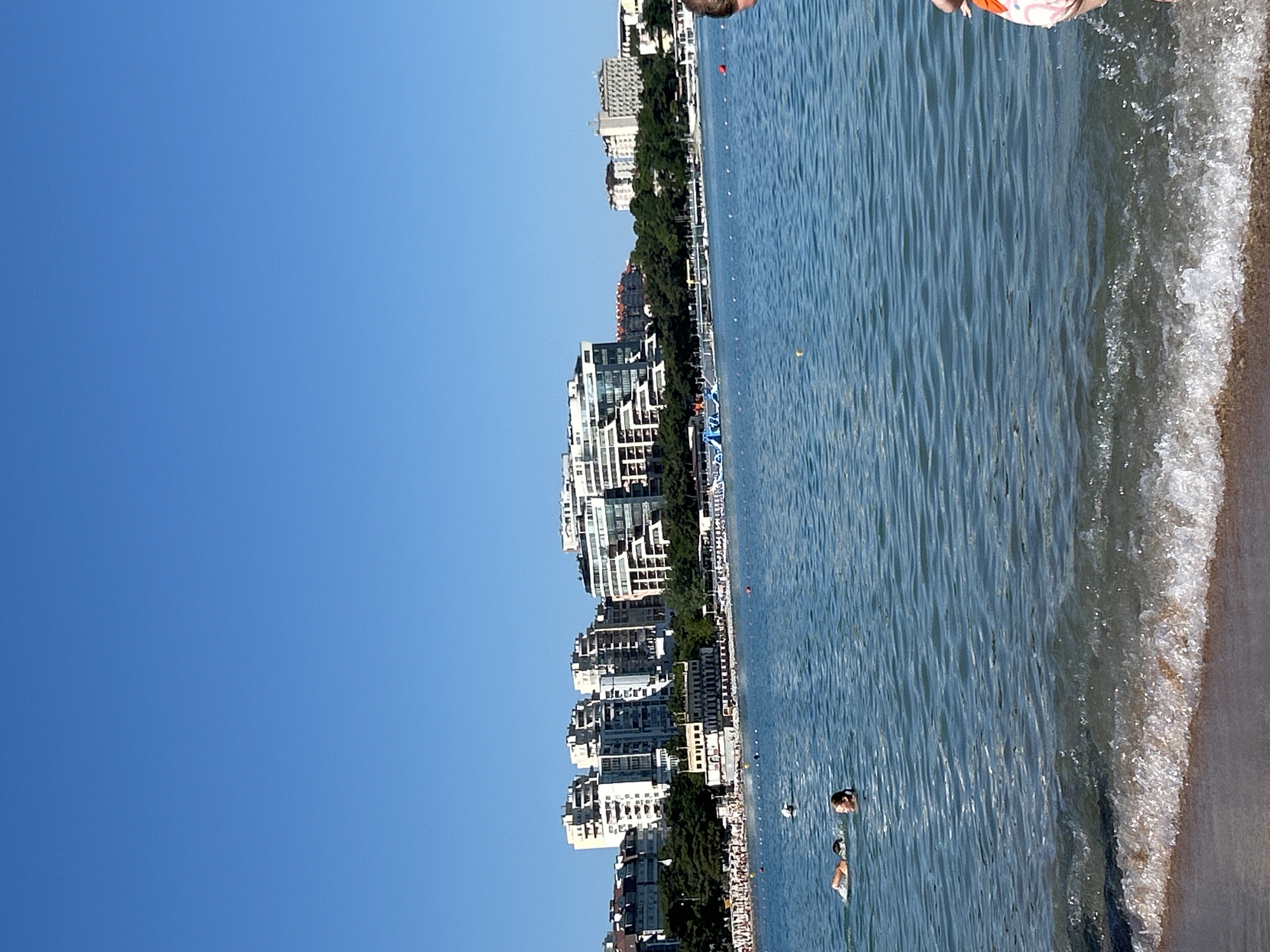 This is a full-frame, eye-level, vertical, medium-shot photograph of the Gelendzhik Bay in Gelendzhik, Russia, on a clear, sunny day. The background features a dense line of trees and several modern, multi-story apartment buildings with balconies, indicating an urban coastal environment. A promenade or beach appears to be situated between the trees and the water, lined with umbrellas and beachgoers.
In the foreground, the clear blue water of the bay is calm with gentle waves lapping at the shore. Two people are visible swimming in the water, about twenty meters from the shore. One person, further away, is swimming face down, while the other, closer to the shore, is swimming face up. In the far right of the frame, a person's legs and lower torso, clad in pink and white patterned shorts, are visible at the edge of the water, suggesting they are either entering or exiting the sea. Several yellow and red buoys are scattered across the water. The sky is a uniform, bright blue with no clouds. The image depicts a typical summer scene at a popular seaside resort.