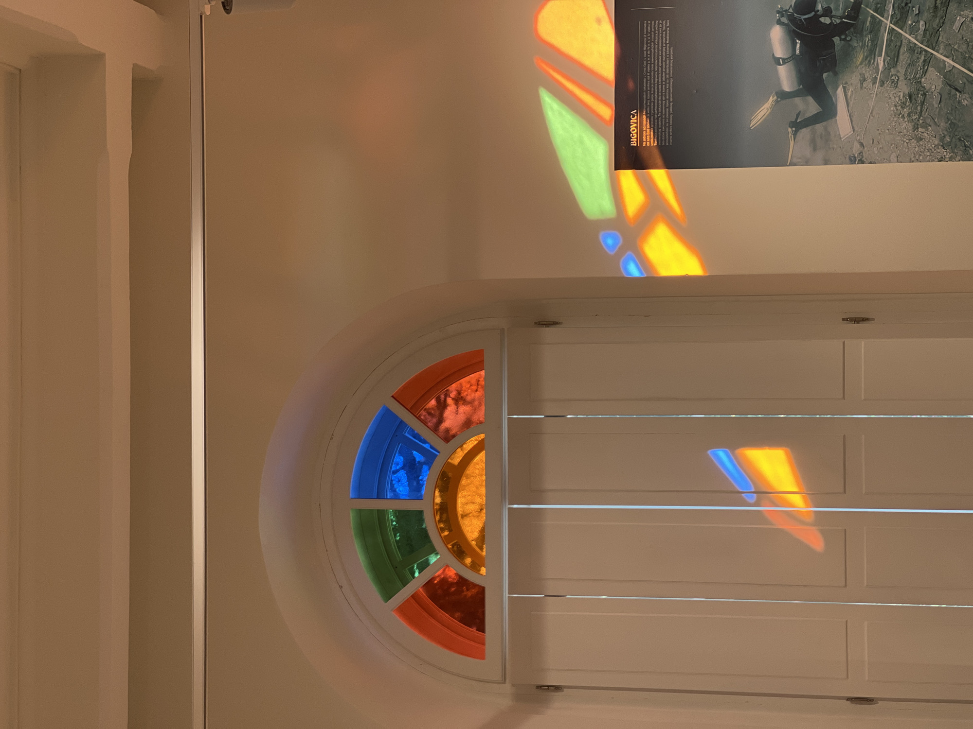 The image captures a section of a wall with a white door and an arched transom window filled with colored glass. Light shines through the colored glass, casting colorful reflections onto the wall and the door. The transom window has panes of orange, yellow, green, and blue glass. The door appears to be a double door with vertical panels. To the right of the door, a poster is mounted on the wall. The poster features text in a language that appears to be Cyrillic, along with an image of a scuba diver. The overall environment is an interior space, possibly a museum or a public building, given the poster. The lighting suggests the presence of natural light coming through the transom window. Without additional information or context, the specific location in the city of Bar, Montenegro cannot be determined from the image alone.