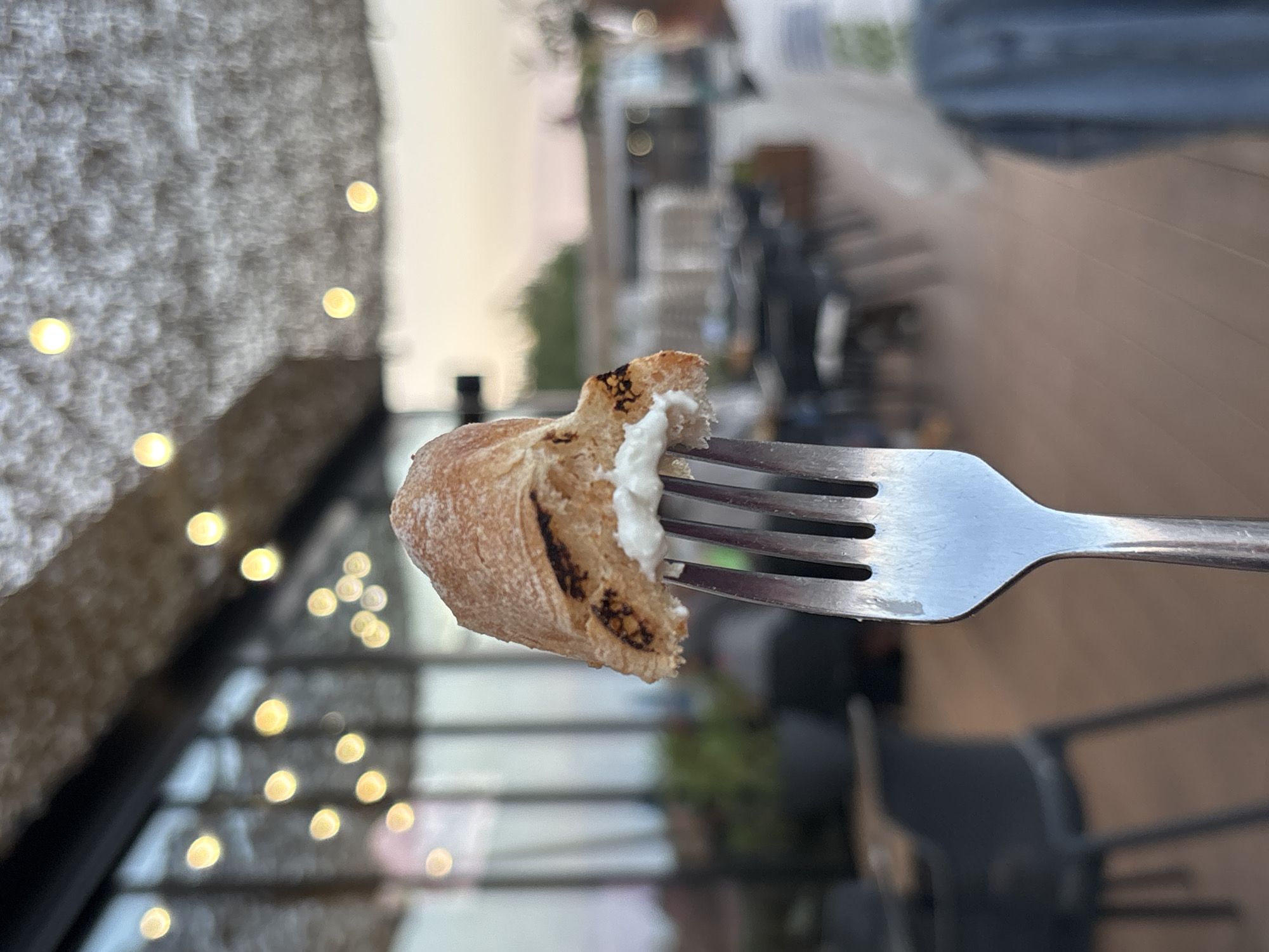 A close-up, eye-level shot captures a piece of toasted bread, approximately 2 inches in length, on a silver four-tined fork, with a dollop of white cream or cheese spread on its underside, positioned centrally in the foreground. The bread exhibits a golden-brown crust with darker, irregularly charred marks. The background is significantly blurred, depicting an upscale outdoor or semi-outdoor dining establishment in Burgas, Bulgaria, during twilight.
Overhead, a light-colored, textured canopy or ceiling structure is adorned with numerous glowing spherical lights, emitting a warm yellow bokeh effect. Behind the bread and fork, blurred dark-colored dining chairs and tables are visible. To the left, large glass panels or windows reveal faint green vegetation and additional ambient lighting. To the right, the soft hues of a twilight sky—pinks, oranges, and light blues—are visible through an open area or panoramic window. A highly blurred figure, wearing a light-colored top and a blue garment, is partially discernible on the far right. The floor appears to be composed of light brown planks or tiles. The scene suggests a relaxed dining atmosphere in a contemporary setting.