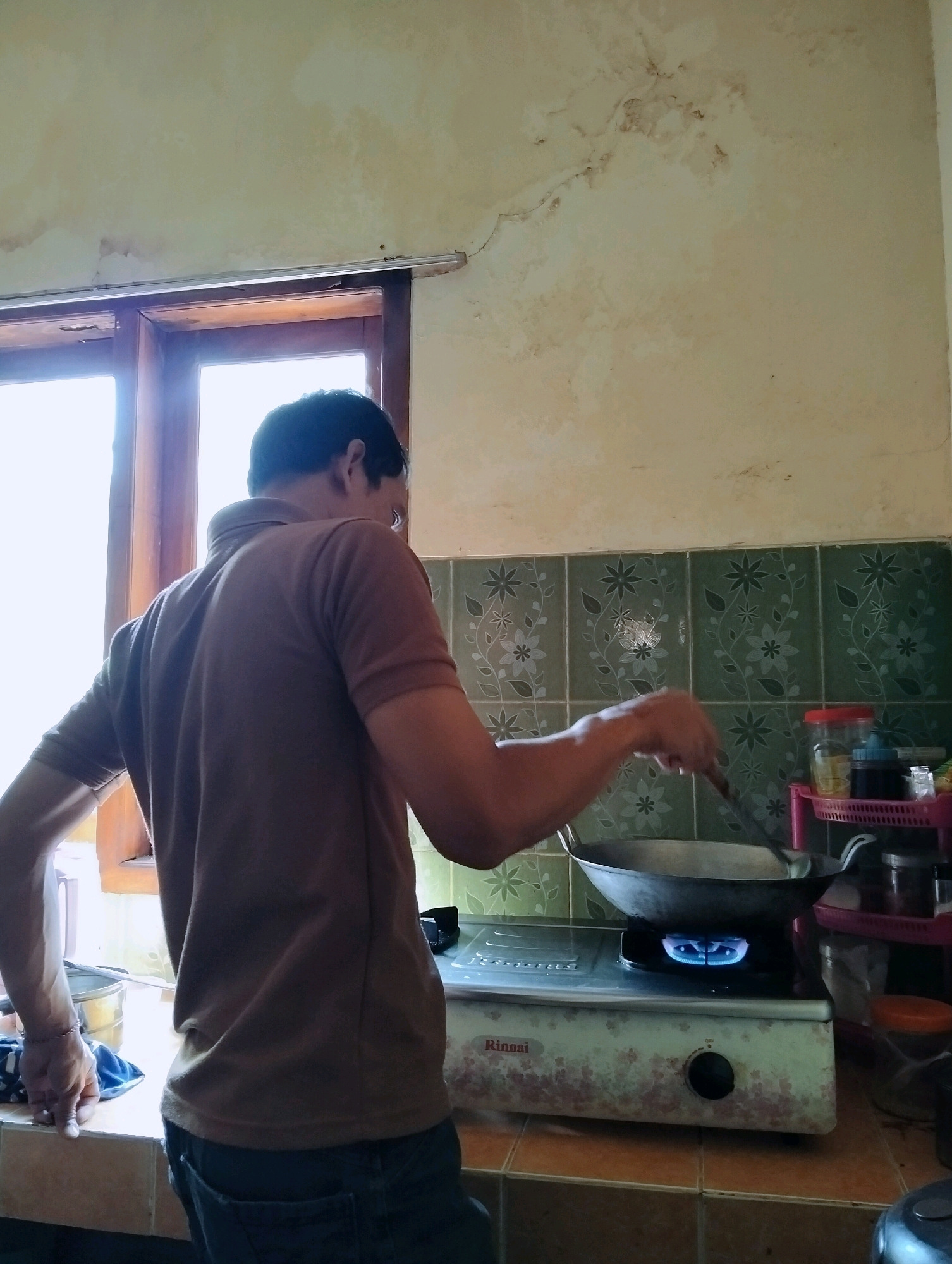 A man stands at a stove in a kitchen, cooking. The man is wearing a brown t-shirt and dark pants. He is holding a spatula in his right hand, stirring something in a wok on the stove. The stove is a Rinnai gas stove with a blue flame underneath the wok. Behind the stove, there are green tiles with a floral pattern. To the right of the stove, there is a pink shelf with jars and bottles on it. The kitchen has a window to the left of the man, letting in natural light. The walls of the kitchen appear to be stained or water-damaged in places. The overall scene suggests a simple, domestic setting in a home. There is no information about the location within the city of Tipar, Indonesia.