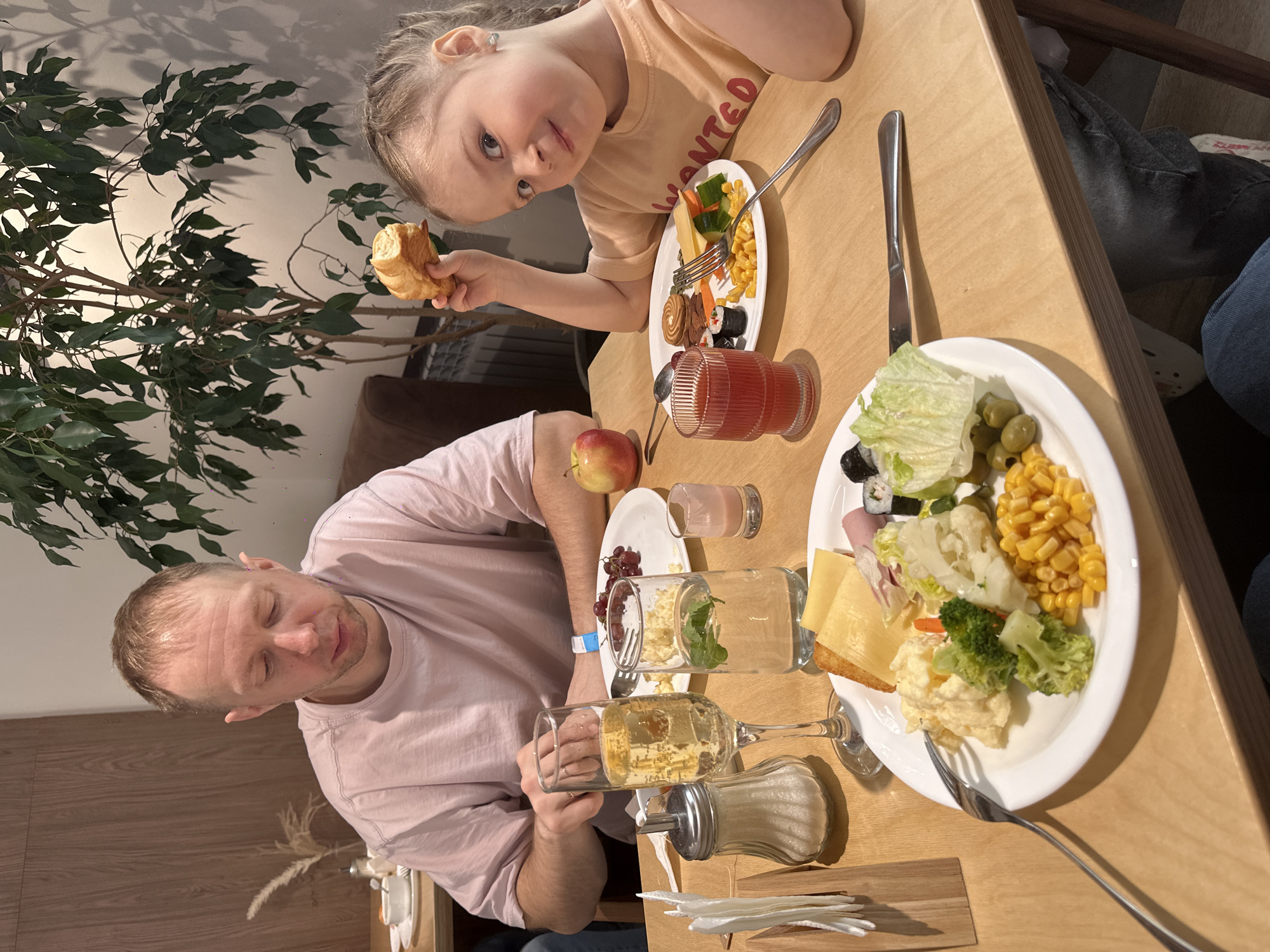 A horizontal, full shot captures a man and a young girl at a wooden table laden with food, presumably dining. The man, with fair skin and short hair, wears a light pink t-shirt. He sits to the left of the frame, holding a glass of amber-colored liquid. Next to him are an apple and several glasses, including one containing a leafy green substance. A sugar dispenser sits near his hands, and plates of food are arranged around him. The young girl, fair-haired with her hair in braids, is positioned to the right, wearing a tan t-shirt with red lettering. She holds a croissant and looks directly at the camera with a curious expression. A plate of food with vegetables and sushi sits in front of her, along with a glass of red juice. The table is filled with an assortment of food items, including a plate with corn, broccoli, cheese, ham, and lettuce. The background includes a plant on the left and glimpses of a wall. While the city of Glazov, Russia, is specified, there are no distinct location markers visible in the image to confirm it.