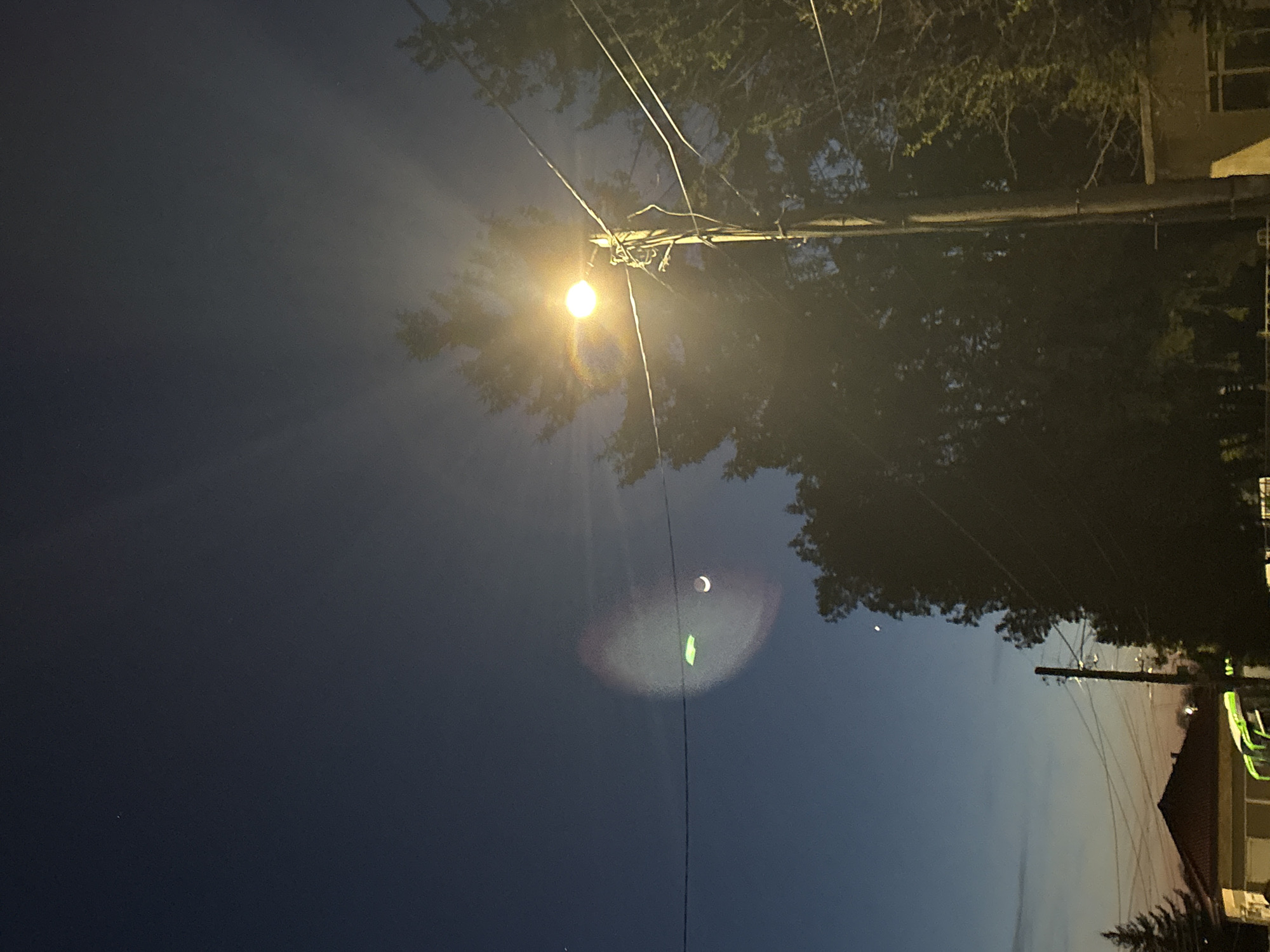 The image shows a night scene in Goritsa, Bulgaria. A street light illuminates the area above, with its light scattering across the sky. A crescent moon is visible in the upper-left portion of the sky, near the light post. Power lines run across the frame, intersecting with the light post. Trees are visible along the top of the frame, obscuring some of the sky. A building is visible on the right-hand side of the frame.