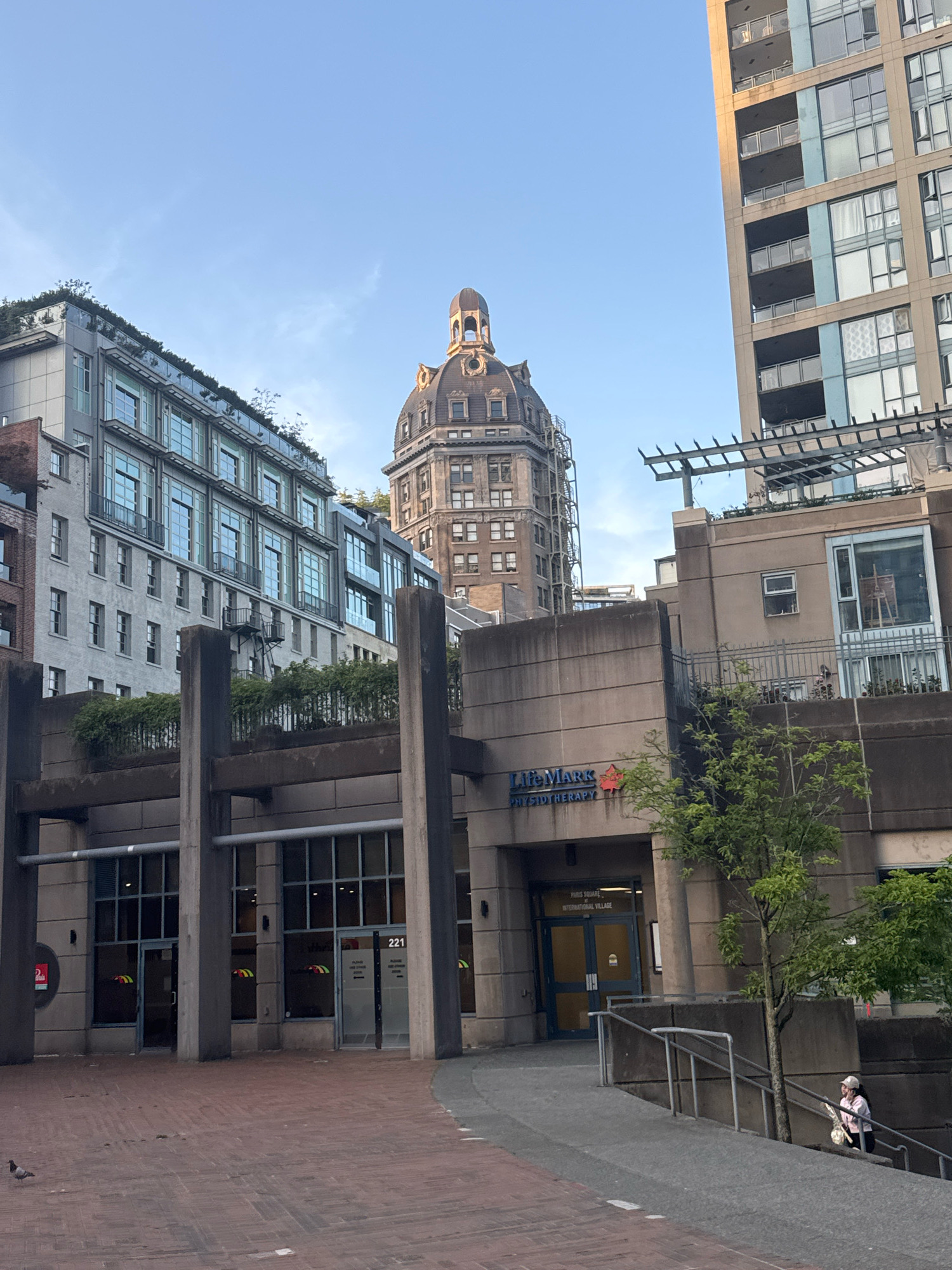 The image depicts an outdoor urban scene with multiple buildings of varying architectural styles under a clear blue sky. In the foreground, a brick-paved plaza with concrete pillars and planters is visible. A single pigeon is present on the plaza. To the left, a modern, grey building with multiple stories and numerous windows is prominent, featuring a green rooftop. To the right, a lighter-colored, tall residential building with balconies and many windows dominates the skyline. In the center, a domed, older-style building rises above the surrounding structures. The middle ground features a beige concrete building with a sign reading "Life Mark PHYSIOTHERAPY". A woman wearing a pink jacket and a light-colored hat is seen walking up a set of stairs to the right of this building. The location is identified as the Paris Square building at 221 West 7th Avenue in Vancouver, Canada, as indicated by signage.