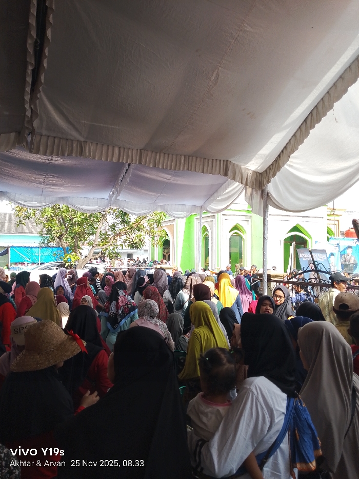 A large crowd gathers under a white tent. The majority of people, primarily women, wear headscarves in various colors. In the background, a vibrant green mosque with arched windows is visible. A tree with sparse foliage stands nearby. Banners and posters can be seen at the rear of the crowd. The scene suggests a community event or religious gathering.