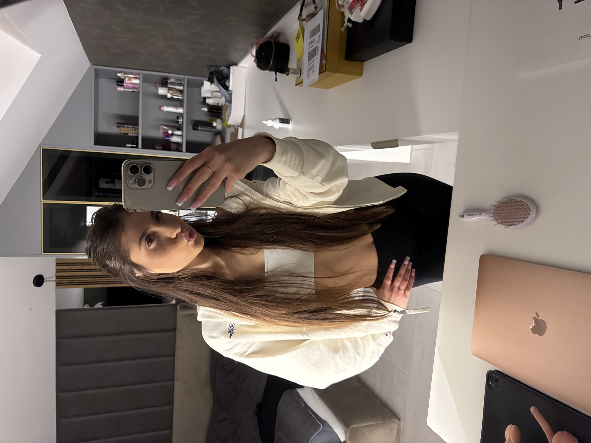 A young woman is taking a selfie in a room. The room includes a vanity with a laptop, tablet, hairbrush, and various cosmetic items on the vanity surface. In the background, a bed with a gray headboard and various shelves of items can be seen. The location is not specified within the image.