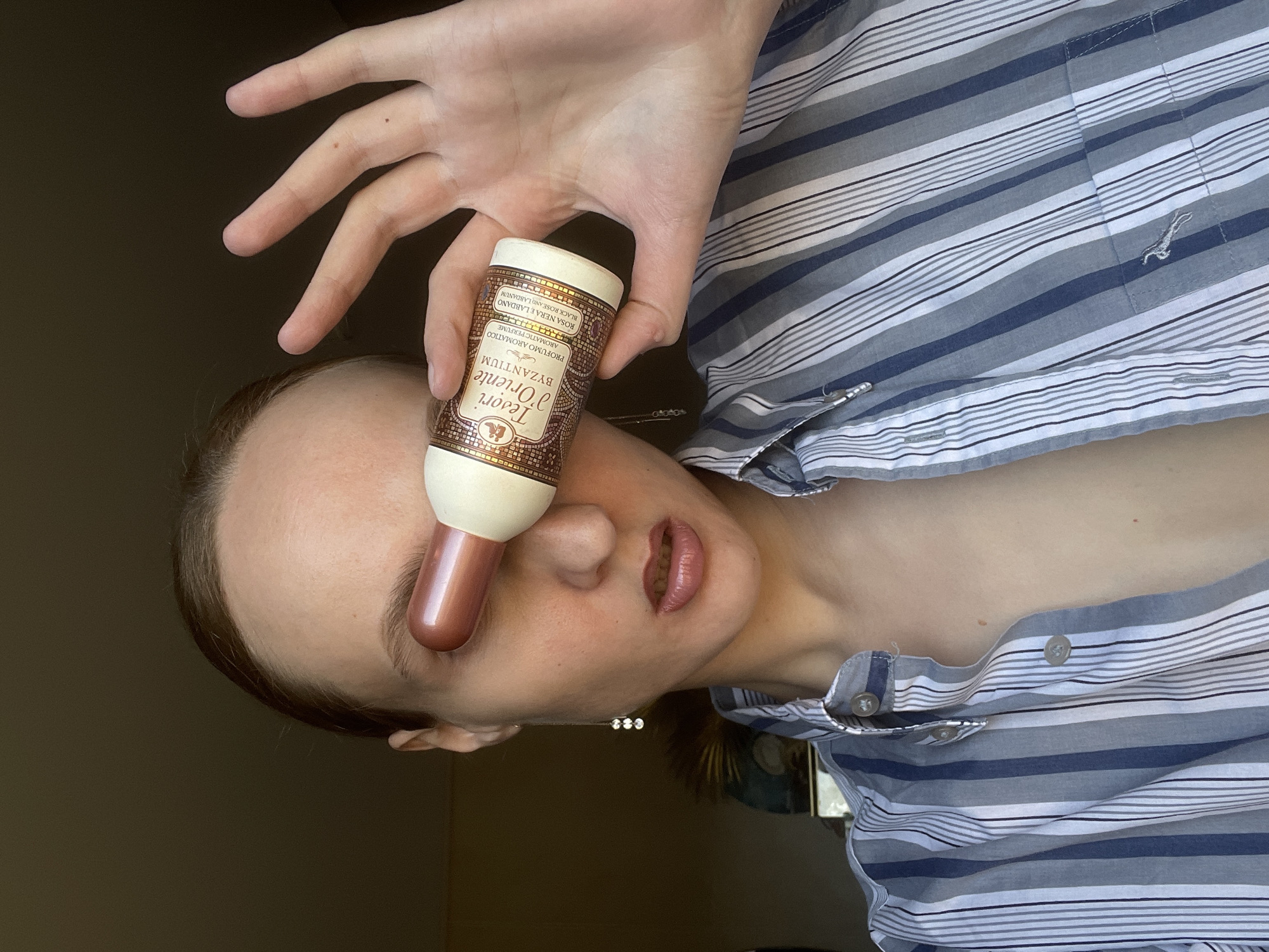 A woman is holding a bottle of Tesori d'Oriente Byzantium perfume near her nose. The bottle is brown and white with text in Italian and English. She is wearing a striped blue and white shirt. Her hand is above the bottle, fingers spread. She has light skin and brown hair. She is wearing a necklace with three small stones. The background is a plain wall. The location is Unknown, Unknown.