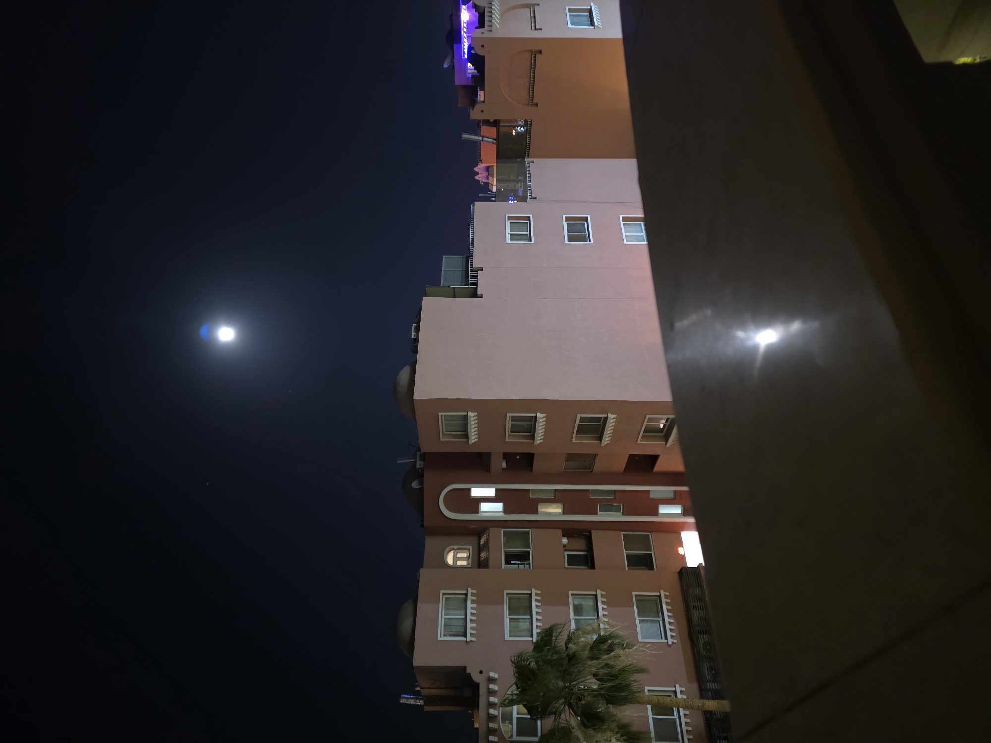 Captured at night, the photograph presents a partial view of a building in Hurghada, Egypt. The structure, painted in varying shades of peach, features a mix of square and arched windows. A palm tree is situated at the building's base. The night sky is visible, punctuated by a bright moon. The building is framed on one side by a dark, shadowed wall.