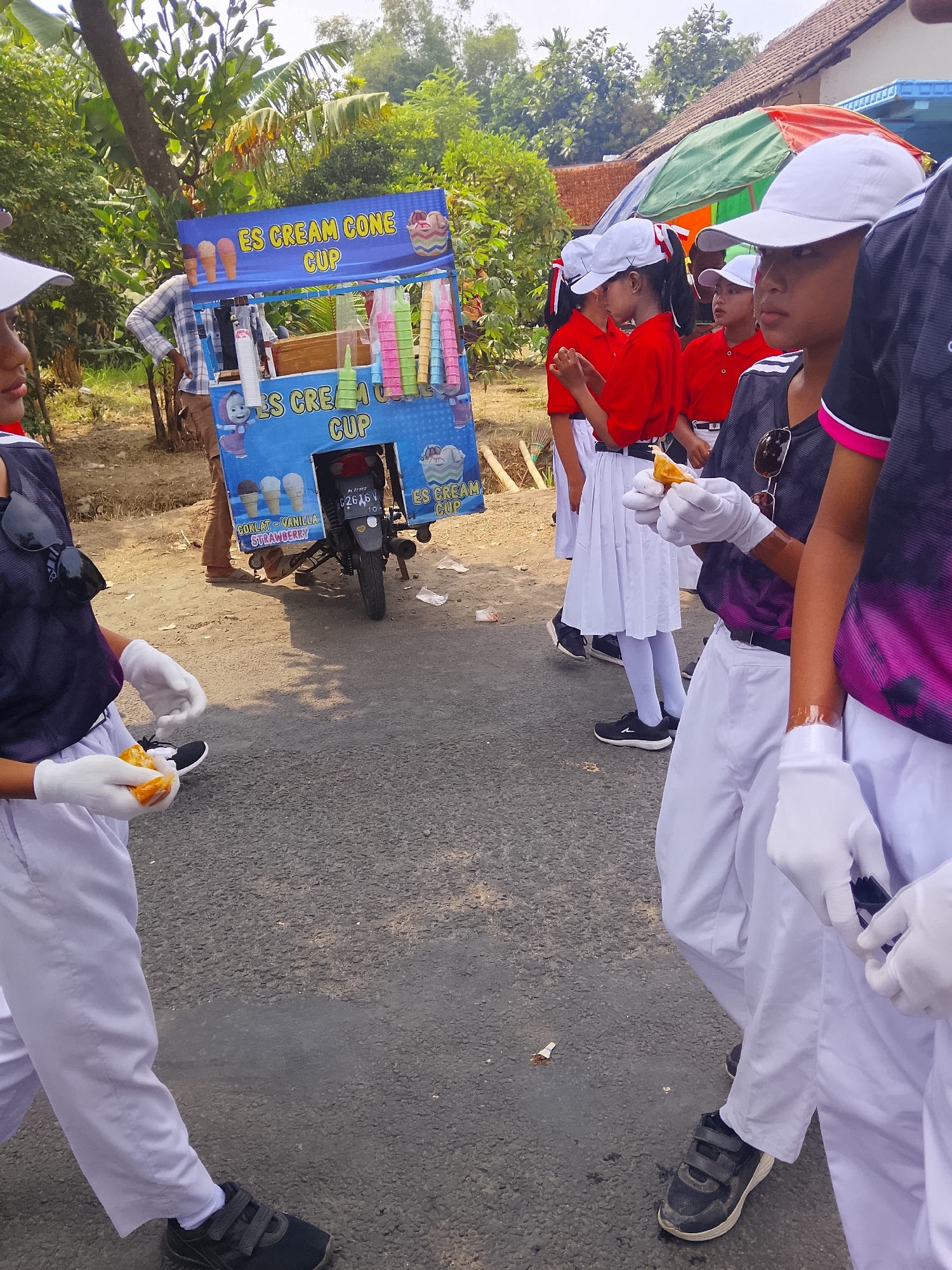 In Bangsri, Indonesia, a group of children, some wearing white caps and red shirts, others in athletic wear and white pants, stand on a paved road near an ice cream cart. The ice cream cart, decorated with images of ice cream cones and cups, is blue and has text indicating "ES CREAM CONE CUP". A motorcycle is parked behind the cart. Some children are wearing white gloves and holding food items. The environment appears to be outdoors, with trees and buildings visible in the background under a bright sky.