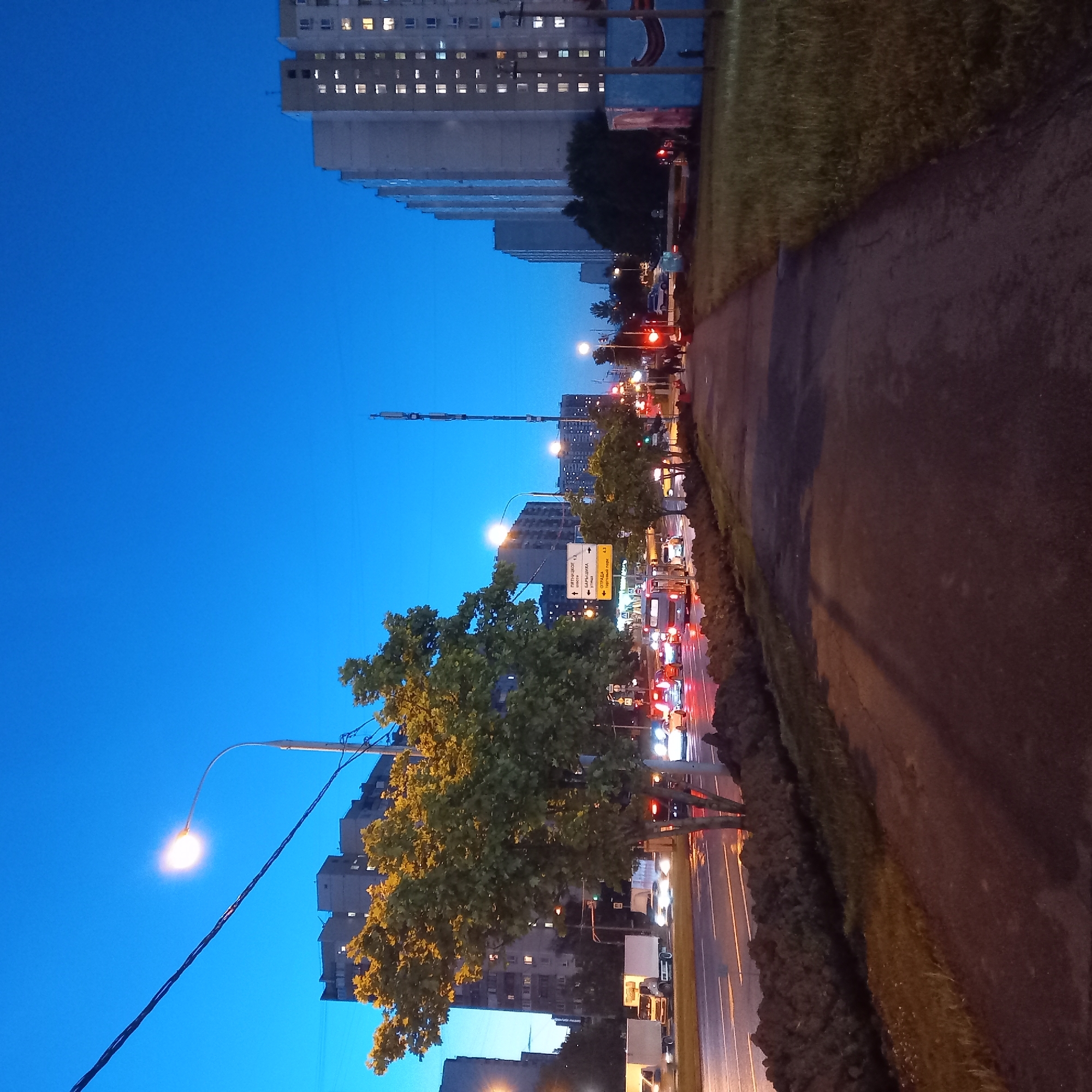 The image depicts a street scene in the city of Khimki, Russia, at dusk or early evening. The sky is a deep blue, indicating the transition from day to night. A brightly lit street lamp is visible in the upper left quadrant, illuminating a power line. On the left side of the frame, several multi-story apartment buildings are silhouetted against the sky, with some windows lit. A large tree with green foliage is prominent in the mid-left.
The street itself is wet, suggesting recent rain, and traffic is present. Several vehicles, including trucks and cars, are moving along the multi-lane road. Headlights and taillights are visible, creating streaks of light due to the long exposure. A pedestrian walkway runs along the right side of the road, separated by a low mound of dark soil or mulch. A few people are walking on the sidewalk in the distance on the right. Further down the road, a tall communication tower is visible. Signage on poles indicates directions to "Пятницкое шоссе" (Pyatnitskoye Highway) and "Отрада" (Otrada). A blue structure with painted art is visible on the far right. The overall atmosphere is urban, with a sense of activity despite the fading light.