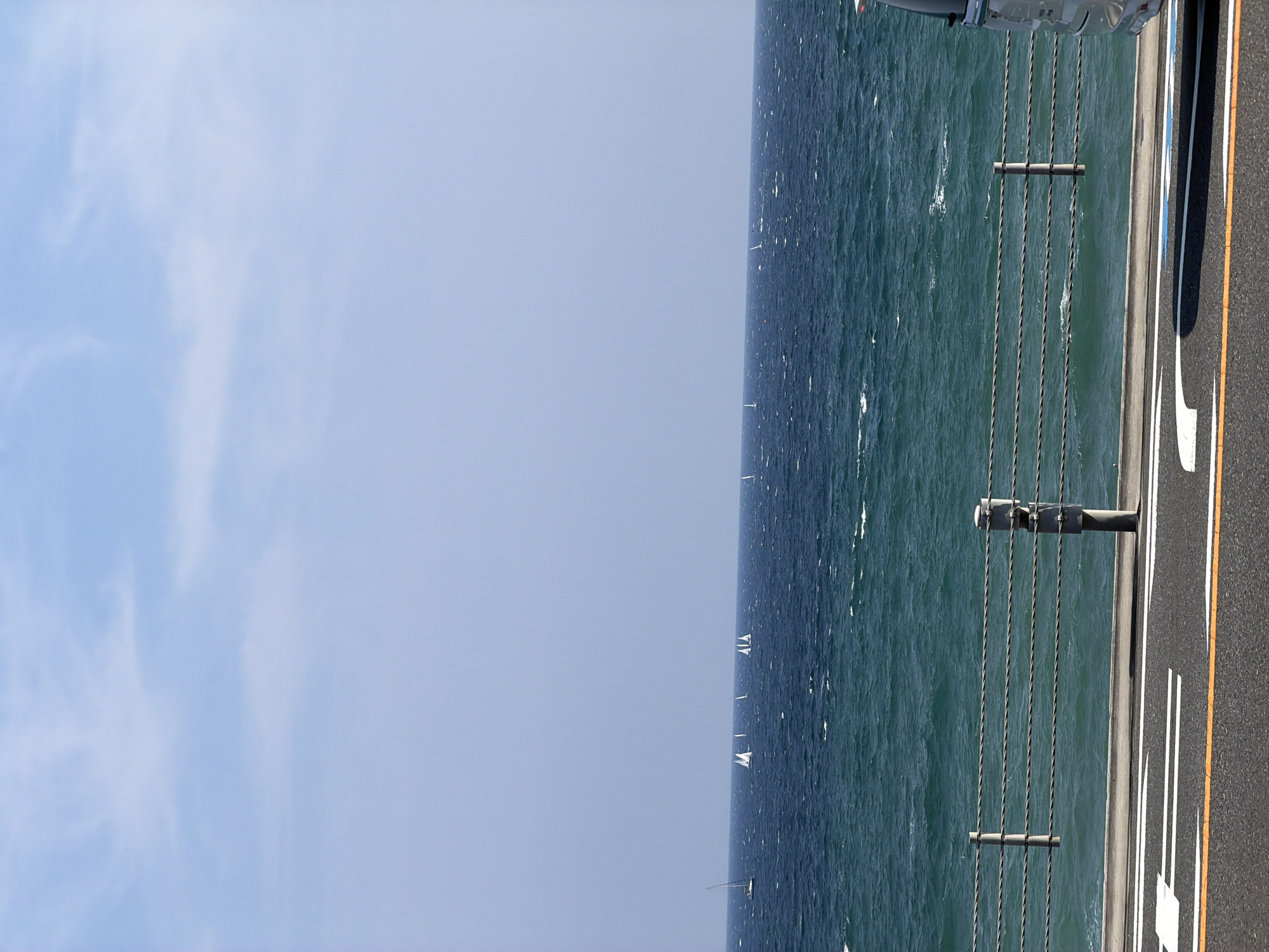 The image captures a view of the Pacific Ocean off the coast of Kamakura, Japan, on a clear, sunny day. The sky is a pale blue with wispy white clouds. The sea is a deep, rich blue-green, with whitecaps indicating moderate wind and wave activity. Several sailboats are visible on the horizon, dotted across the expanse of water.
In the foreground, a paved road with white and orange lane markings runs horizontally across the bottom of the frame. To the left of the road, a metal guardrail with multiple horizontal cables, supported by metal posts, separates the road from the ocean. The guardrail is casting a shadow onto the road. On the far right edge of the image, the side of a silver vehicle is partially visible, suggesting the photo may have been taken from inside or next to a vehicle. The overall impression is one of a scenic coastal location, likely a promenade or roadside viewpoint overlooking the sea.