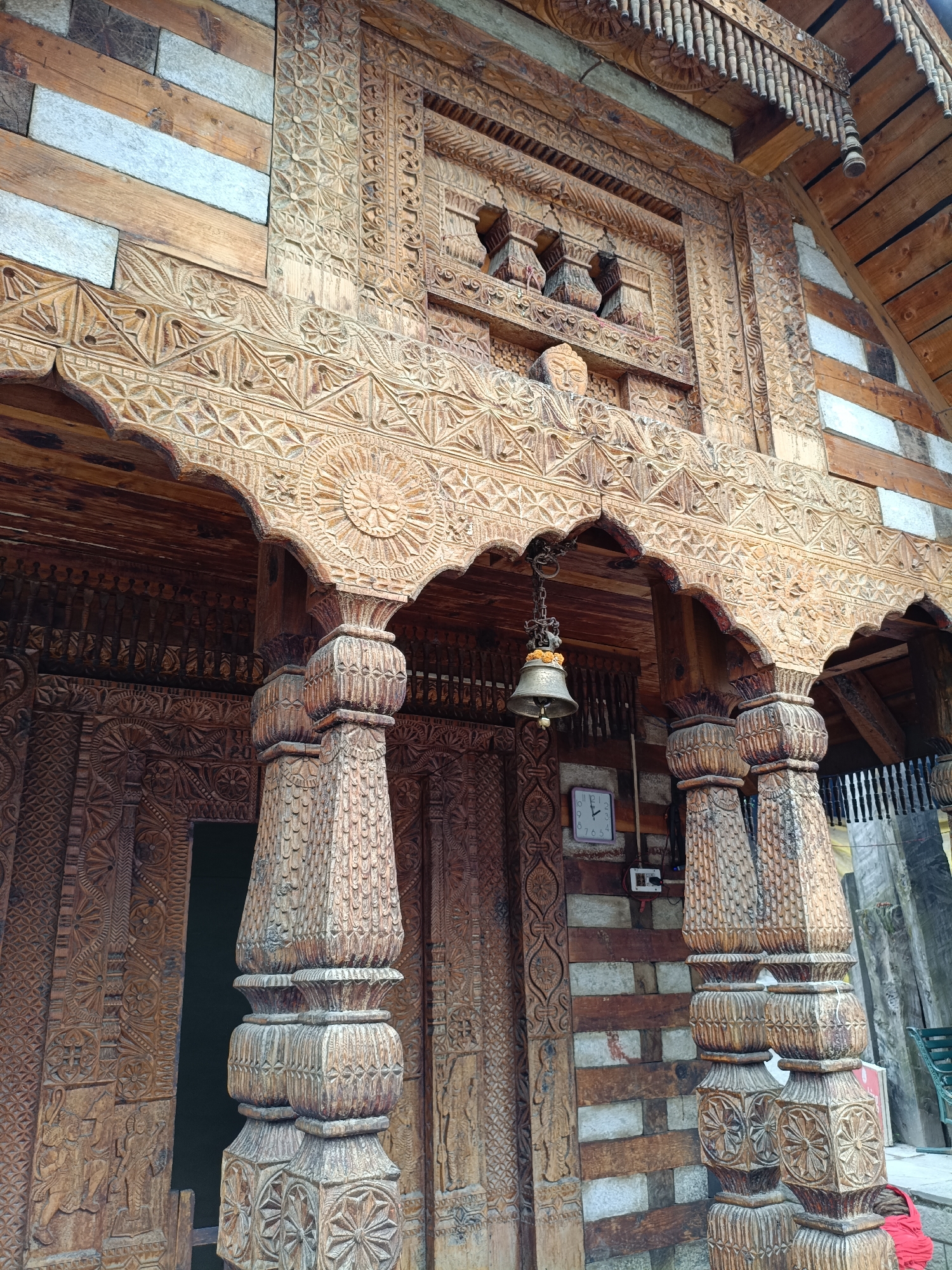 This image captures a close-up view of the intricately carved wooden entrance to a building, likely a temple or traditional house, in Vashisht, India. The architecture features extensive wood carving with geometric patterns, floral motifs, and stylized figures. Several wooden pillars, also ornately carved, support the overhanging roof structure. A brass bell hangs from a chain above an entranceway, and a white clock with black hands is mounted on the wall to the right of the doorway. The building's exterior is constructed from a combination of carved wood and stone or slate elements. The overall scene is characterized by detailed craftsmanship and a rustic aesthetic, indicative of traditional North Indian architectural styles found in mountain regions.
