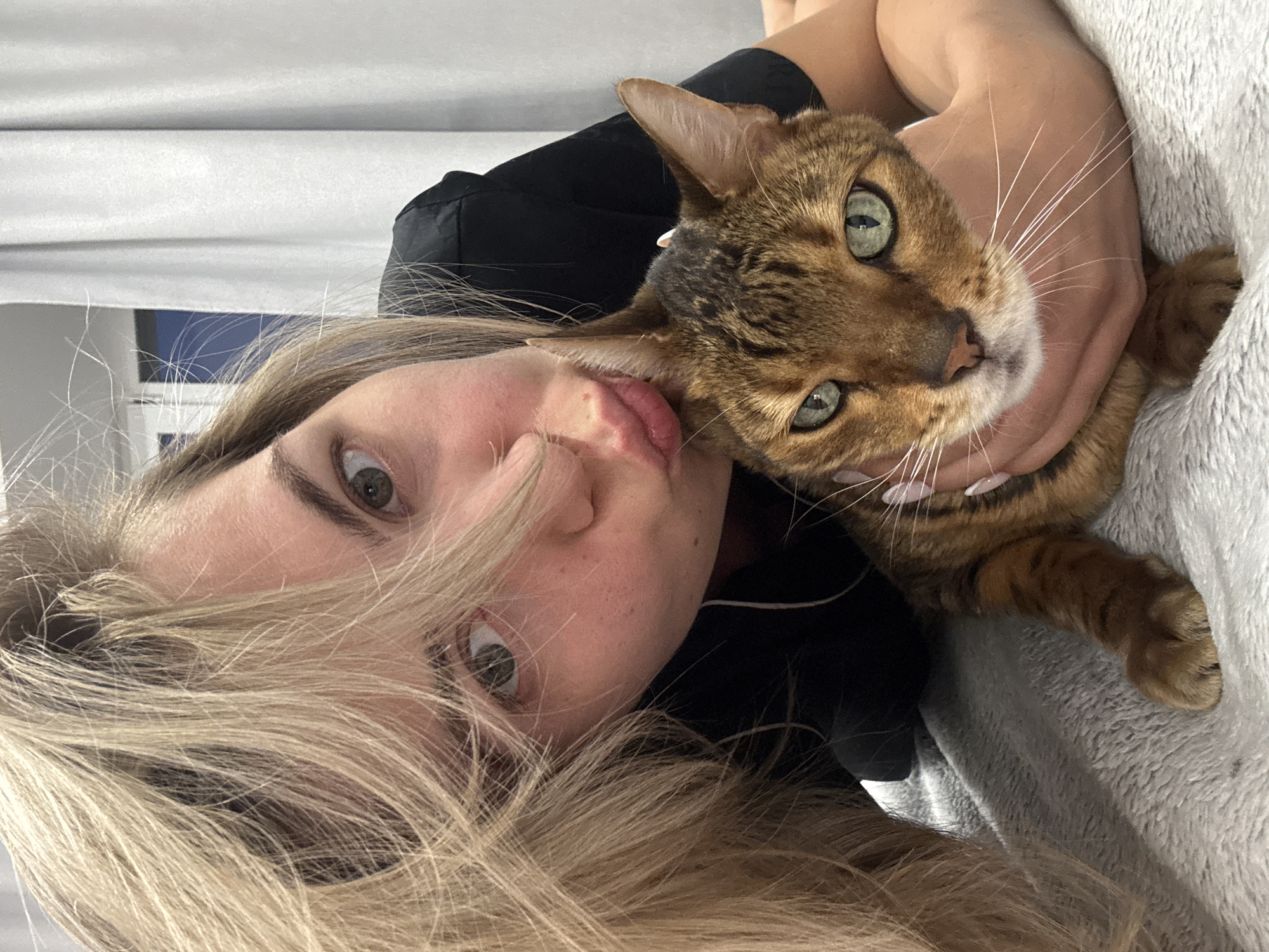 A selfie showing a woman holding a Bengal cat. The woman has long blonde hair and is wearing a black garment. She is making a kissing face towards the cat. The cat is brown with black spots and has green eyes. They appear to be indoors against a gray and white background. The city location is Unknown, Unknown.