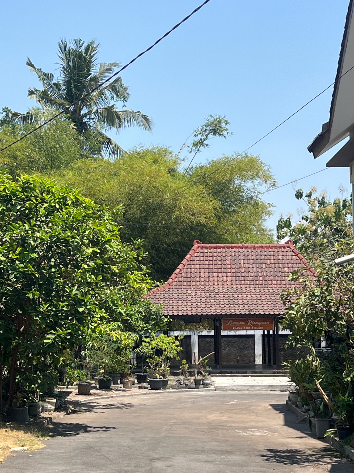 The image shows a building with a red tiled roof, surrounded by lush greenery and trees. In the foreground, there is a paved pathway leading towards the building. The building appears to be a traditional Indonesian structure with an open-air design. A wooden sign above the entrance reads "Limesan Plerengan" and "BUANA ASRI VILLAGE". The environment is tropical, with palm trees and bamboo visible. The sky is clear blue. The image appears to be taken in a rural or semi-rural setting, possibly a village or a resort. The location is identified as Sleman, Indonesia.