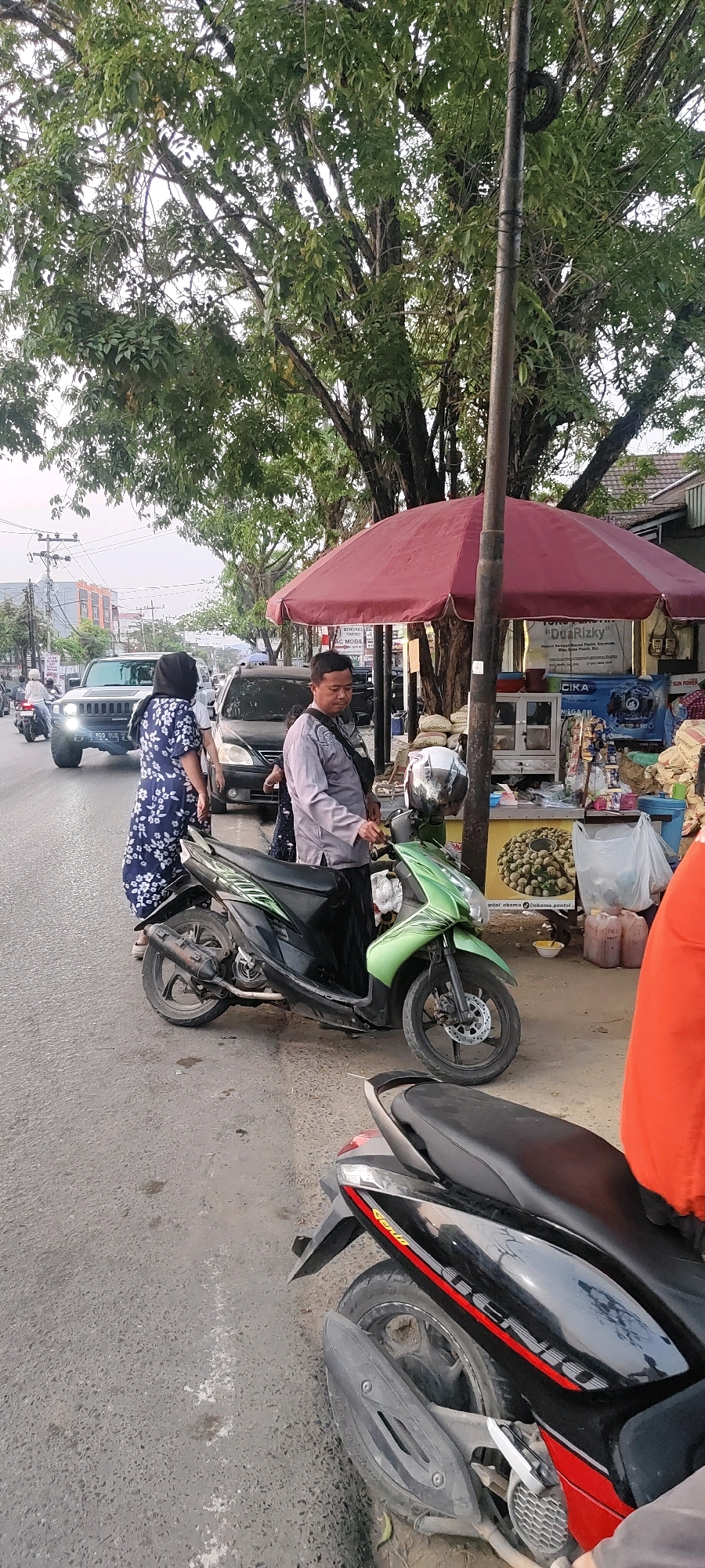 This image captures a street scene in Banjarmasin, Indonesia, likely on a roadside or sidewalk. A man in a grey collared shirt and black pants, possibly a vendor or a passerby, stands next to a green and black motorcycle. He is looking down, seemingly engaged with something near the motorcycle. A woman in a blue and white floral dress and a black headscarf is standing to the left of the motorcycle, also looking downwards. Behind them, there is a stall with a red umbrella and various goods displayed, including what appears to be produce. A black car and a white SUV are visible on the road in the background, along with other vehicles and pedestrians. Trees with dense foliage line the street, providing shade. The overall environment suggests an urban commercial area, possibly a market or a row of shops.