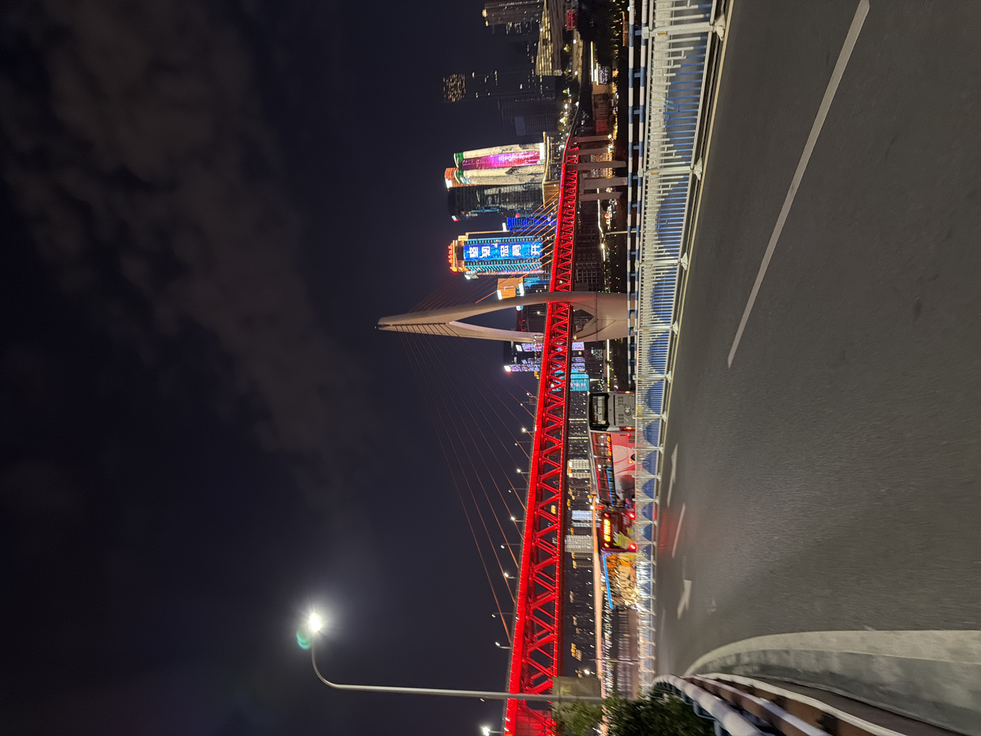 The image captures a night scene in Yuzhong District, China, showcasing a vibrant urban landscape dominated by a brightly illuminated red bridge. The bridge is a cable-stayed structure, with its cables stretching from a tall, angular pylon. The deck of the bridge is lined with red lights, creating a striking visual effect. In the background, a cluster of modern skyscrapers, also adorned with colorful lights, punctuate the dark night sky. The lower portion of the image shows a paved road with white lane markings, indicating it is likely a thoroughfare. A white and blue striped barrier runs along the edge of the road. Two buses are visible on the bridge, one with a yellow and orange livery and the other with a predominantly white and pink design. The overall atmosphere is one of a bustling city at night, characterized by impressive infrastructure and illuminated architecture.