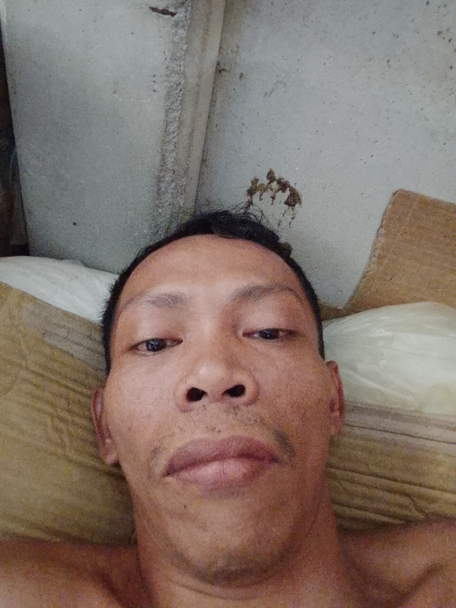 A close-up selfie depicts a bare-chested adult male with short dark hair and a prominent nose. His expression is neutral. The background consists of a white pillow, a piece of cardboard, and a concrete wall. The picture was taken in an interior location. There is no verifiable evidence to support the picture was taken in Bukit Ketok, Indonesia.