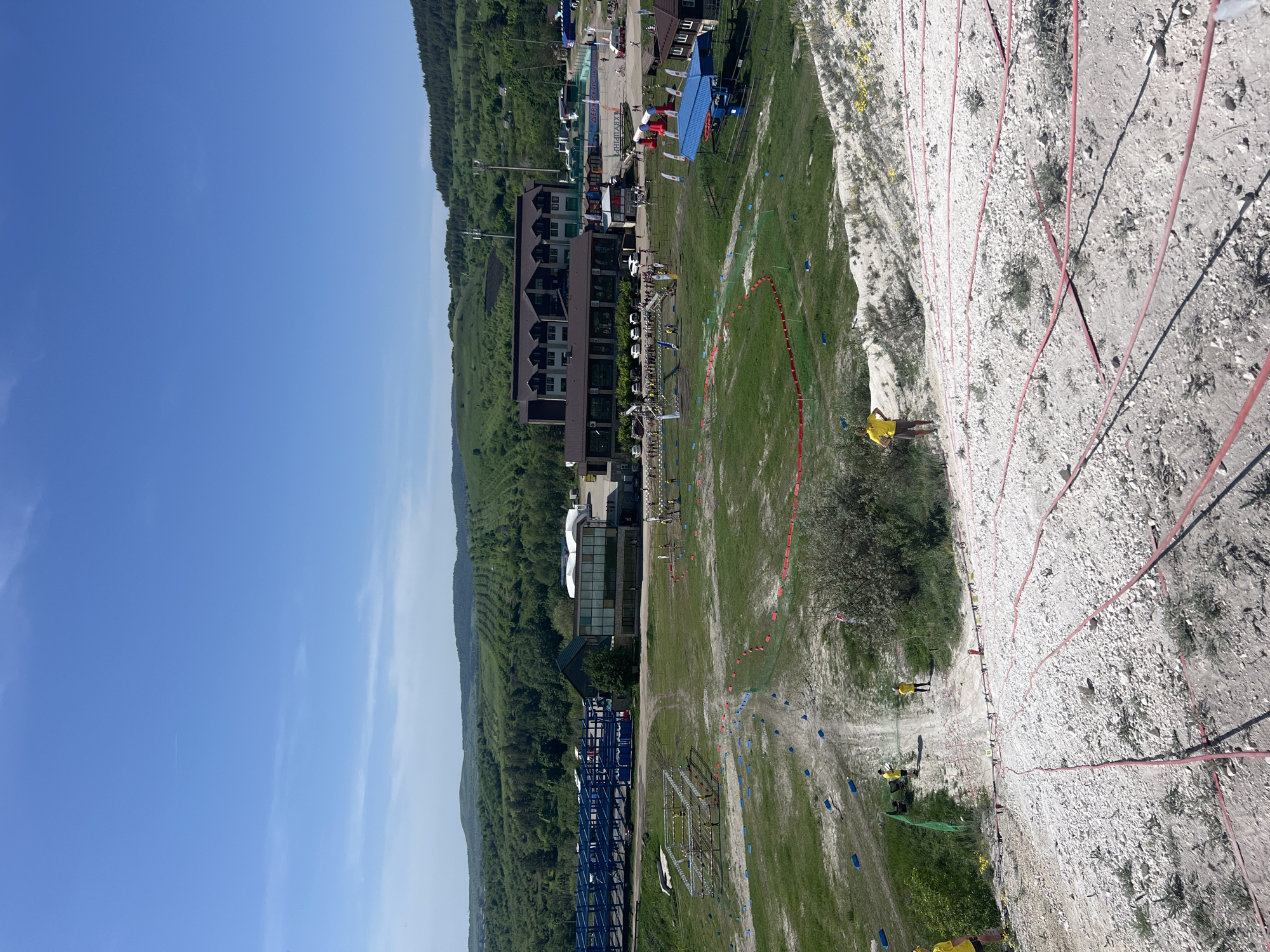 The image depicts a landscape in Khvalynsk, Russia. To the left, there is a building with a modern architectural style, situated amidst a green forested area. In the center is a grassy field, with several small objects scattered across it, possibly indicating a sporting event or similar outdoor activity. To the right, there is a steep, rocky slope that appears to be a natural cliff face. The sky above is clear and blue. There are people visible in the lower center of the image. Red markers appear to be set into the cliff face.