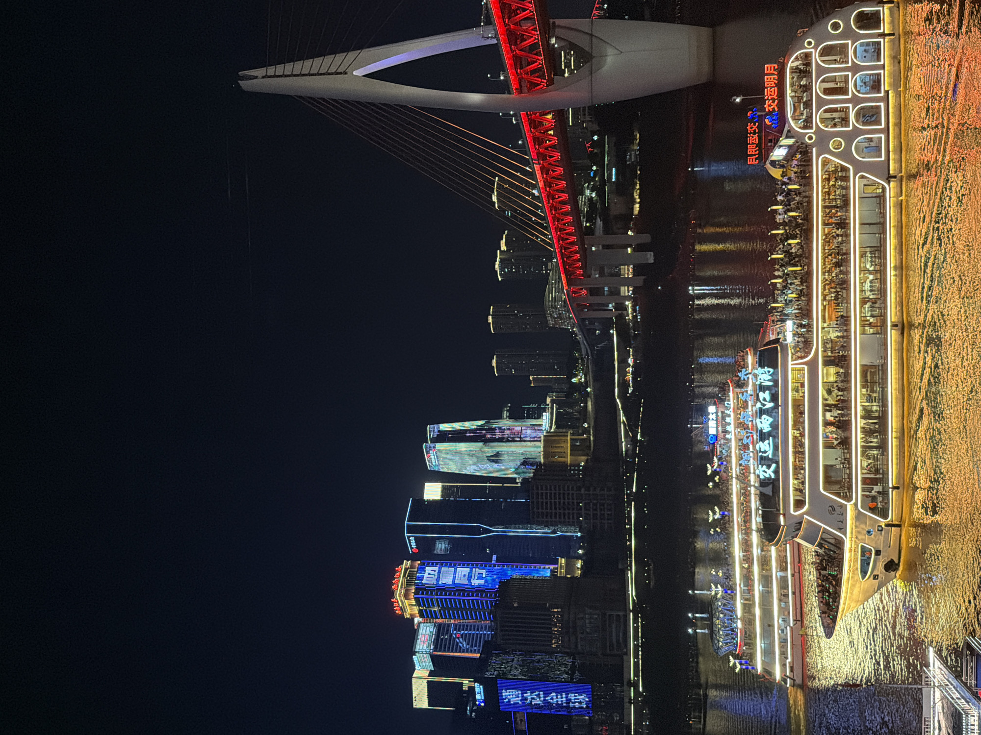 This is a nighttime long-shot photograph of the Yangtze River in the Yuzhong District of Chongqing, China. The scene is dominated by the illuminated Hongyadong Bridge, which is lit with bright red lights. In the foreground, two large passenger boats are navigating the river, their decks crowded with people. The boats are also brightly illuminated with white and yellow lights, creating a golden reflection on the water. Behind the boats, the Chongqing skyline is visible, featuring numerous tall buildings adorned with colorful lights and digital displays. The sky above is dark and clear.