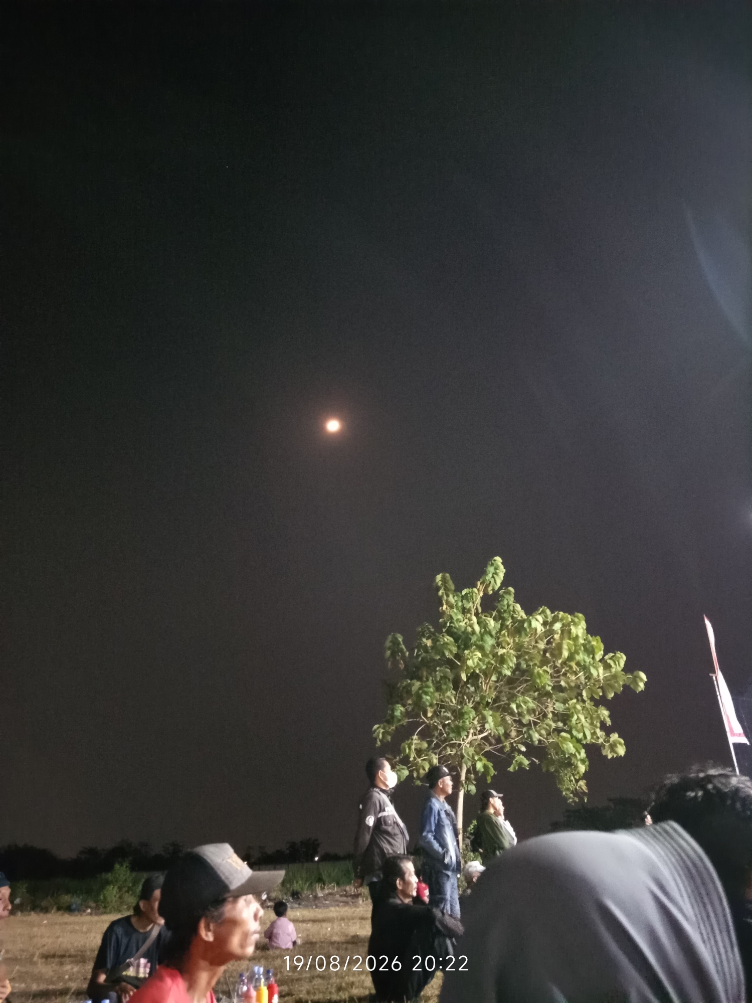 In Canggu, Indonesia, a group of individuals gathers outdoors at night under a dark sky illuminated by a bright, distant light, possibly the moon or a strong lamp. The scene appears to be set in an open, grassy field, with a tree providing a focal point to the right of the frame. Several people are visible, some sitting on the ground, while others stand. They are dressed in casual clothing. In the foreground, a person wearing a baseball cap is seated, looking upwards. In the middle ground, several men are standing near the tree. A child is also visible in the background, seated. The overall atmosphere is relaxed and informal, suggesting a communal gathering or event taking place after dark. The timestamp in the lower right corner indicates the image was captured on August 19, 2026, at 20:22.