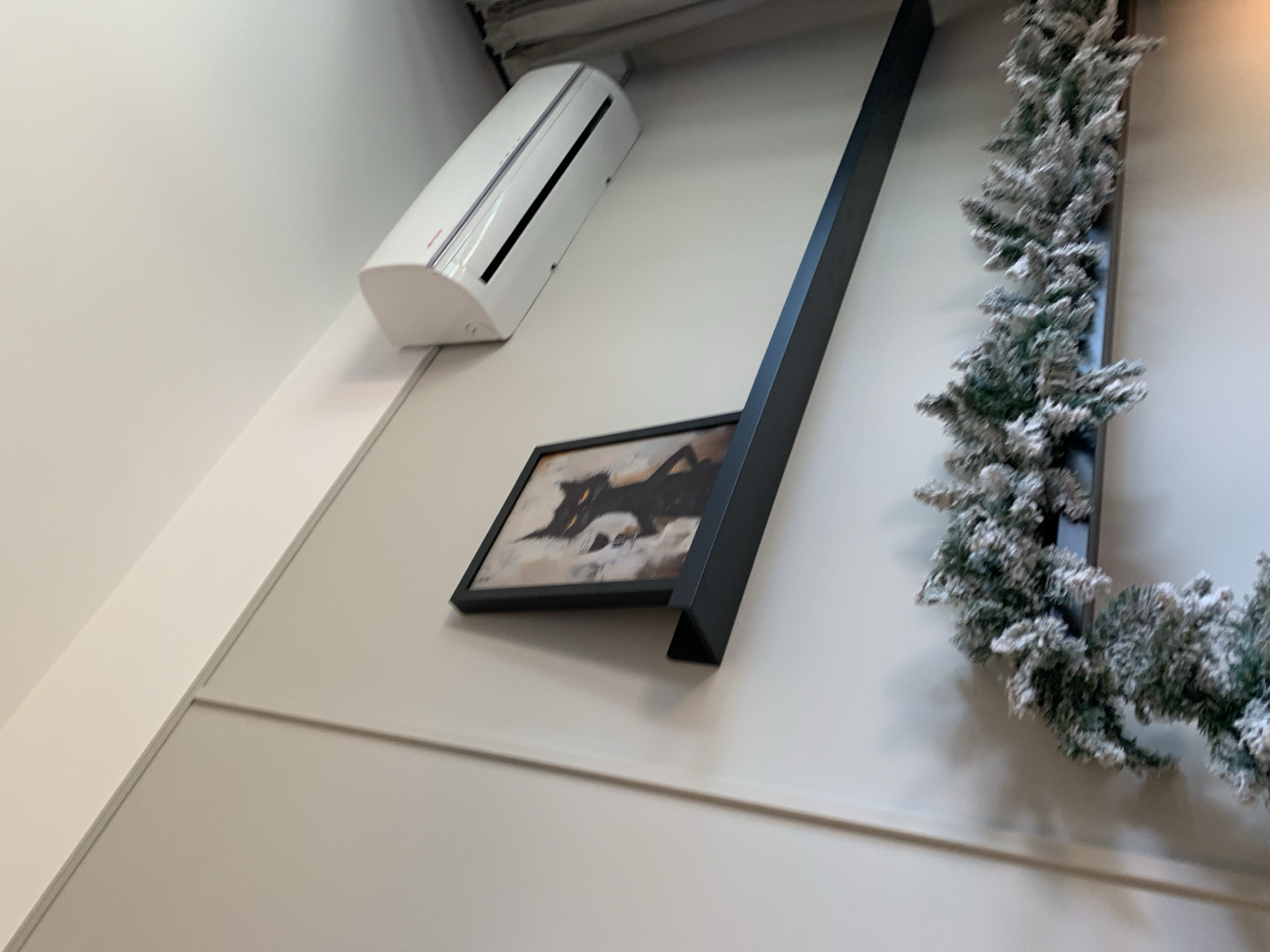 The image shows an indoor space with a white wall. Mounted on the wall are an air conditioning unit, a framed picture, a black rectangular object, and a decorative garland resembling a Christmas wreath. The picture appears to be a modern abstract artwork. The environment seems to be a modern interior. There is no specific information in the image to identify the location as Belgrade, Serbia.