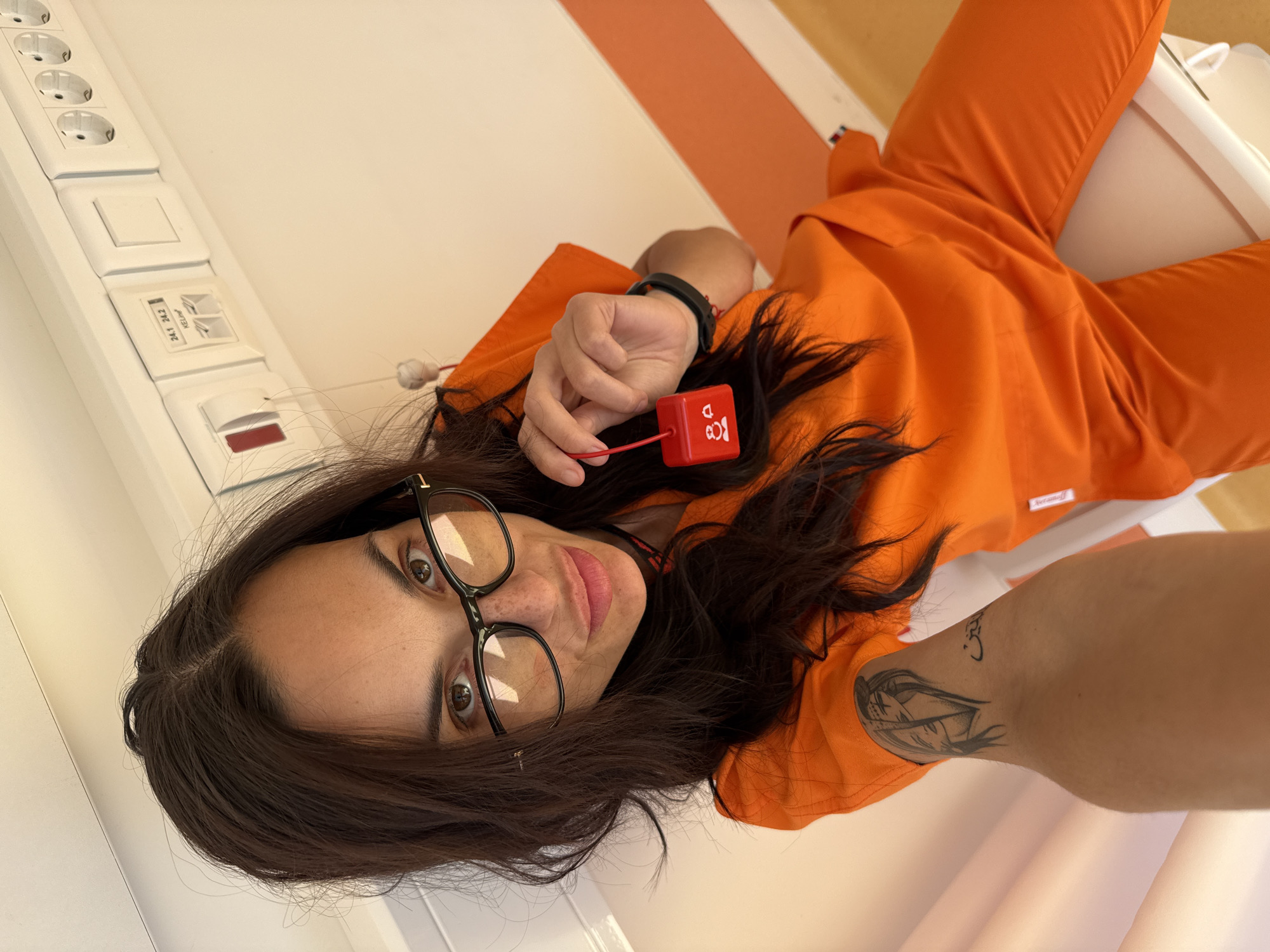 A woman wearing glasses and orange clothing is lying on a white surface. She holds a red device in her hand. A tattoo is visible on her arm. A white wall with electrical outlets is nearby. The scene is indoors. Location information is not discernible.
